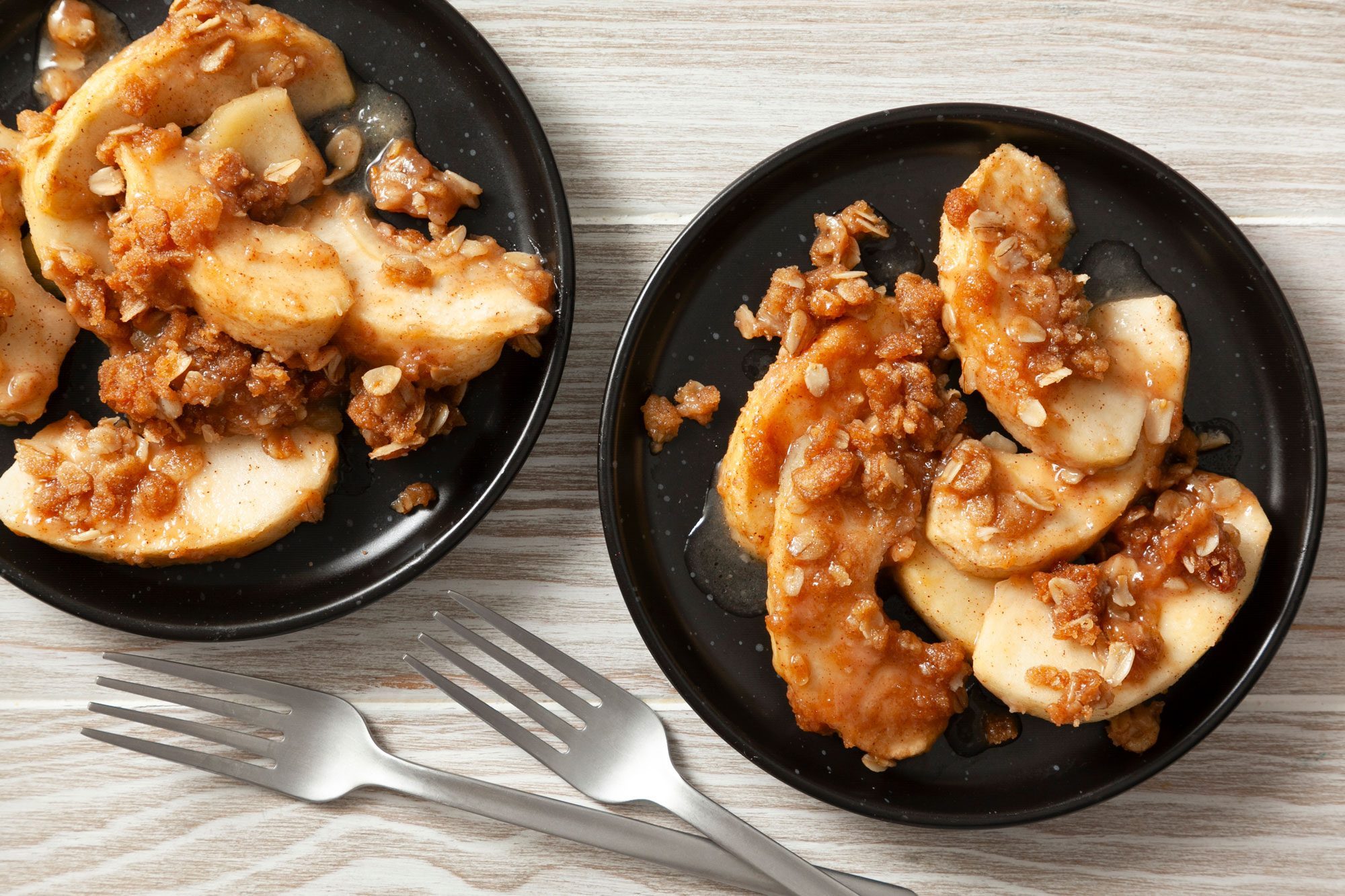 overhead shot; wooden background; Vegan Apple Crisp served in two small black plates and with two silver forks;