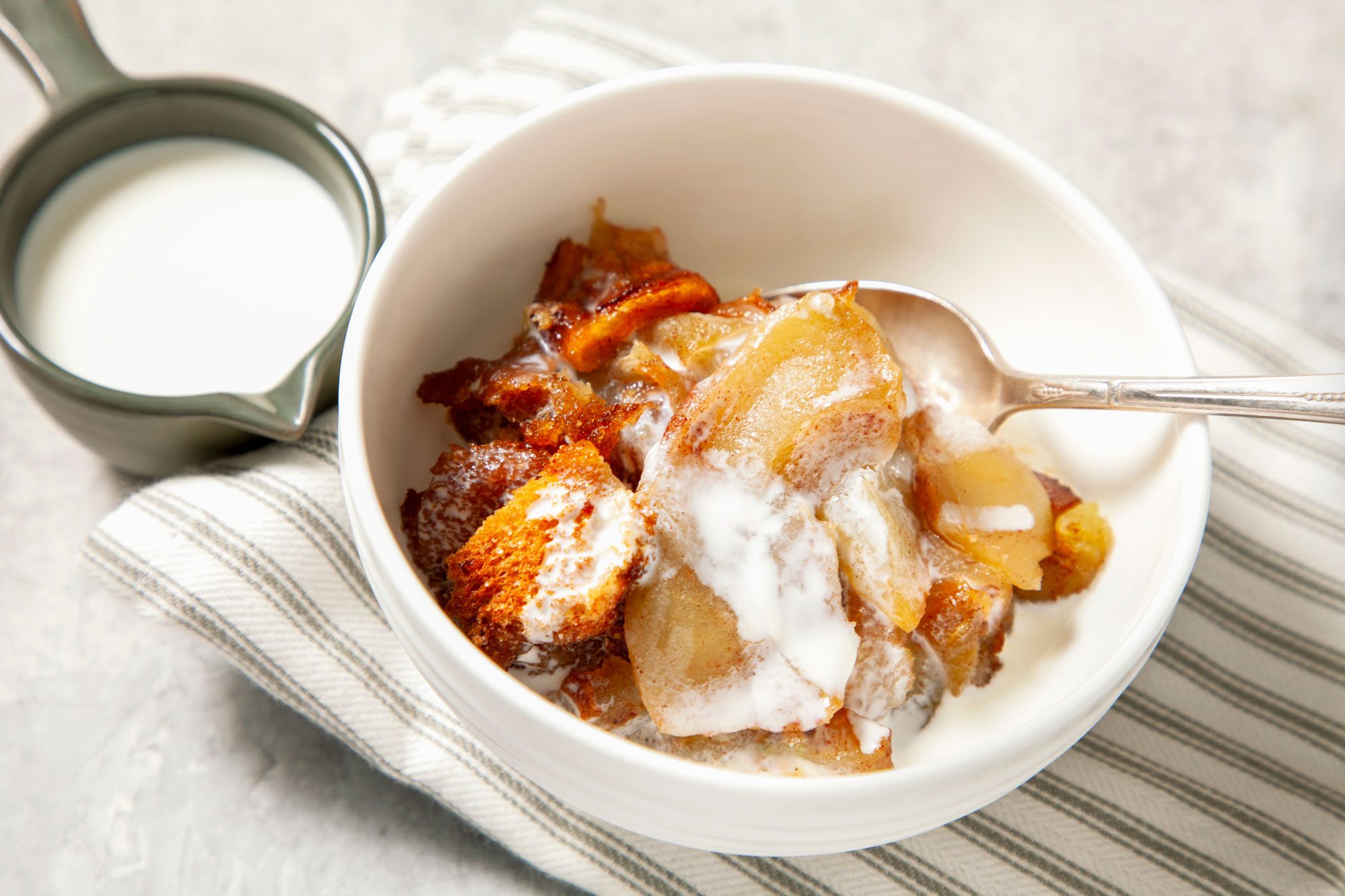 Close shot of Apple Brown Betty; served warm with cream in bowl; spoon; napkin; grey background;