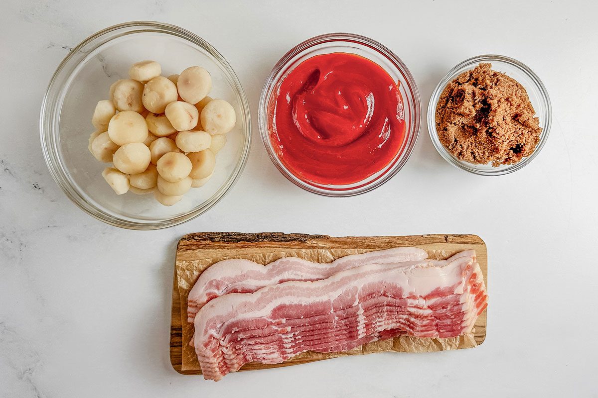 Ingredients for Taste of Home bacon wrapped water chestnuts in glass bowls and a wooden board on a marble surface.