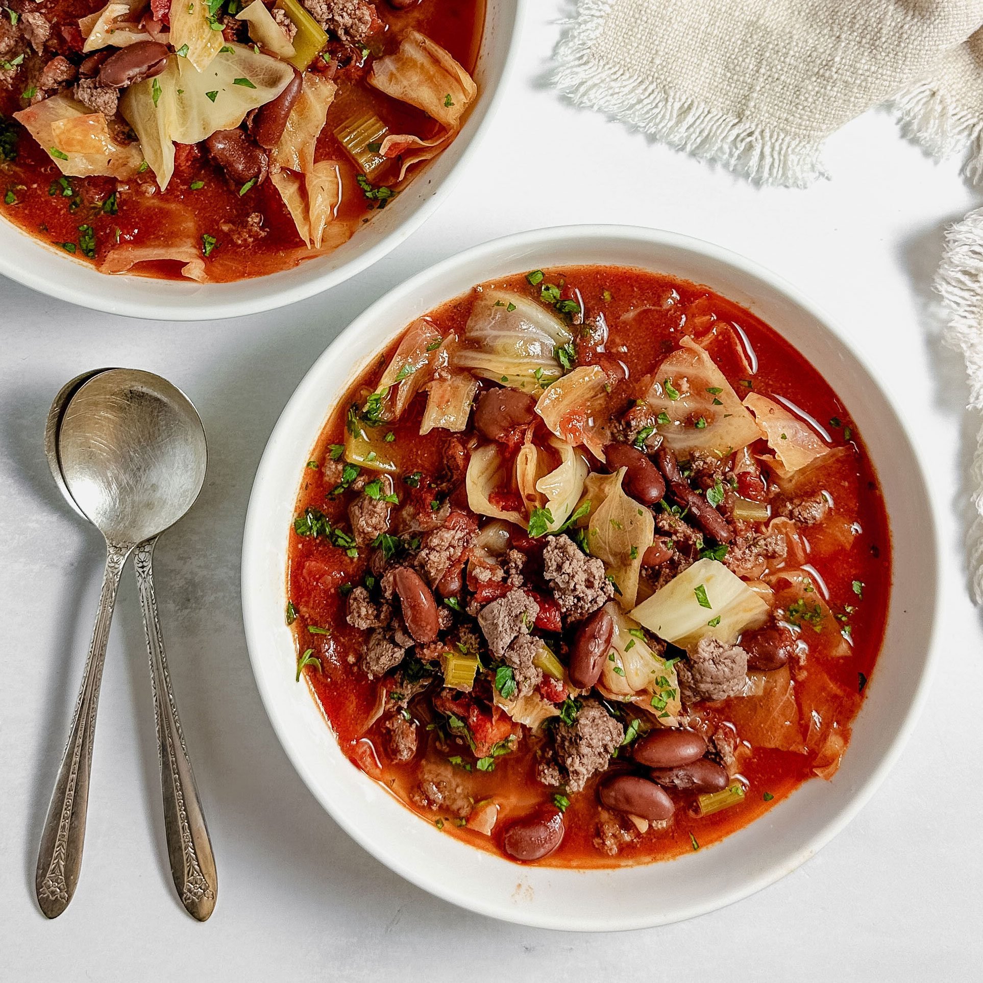 Taste of Home Beef Cabbage Soup in white ceramic bowls with silver spoons and a linen napkin on a marble surface.