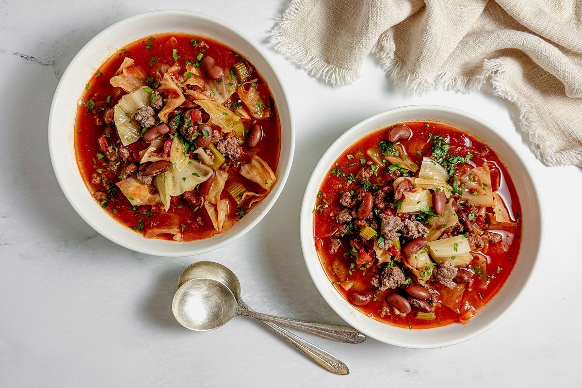 Taste of Home Beef Cabbage Soup in white ceramic bowls with silver spoons and a linen napkin on a marble surface.