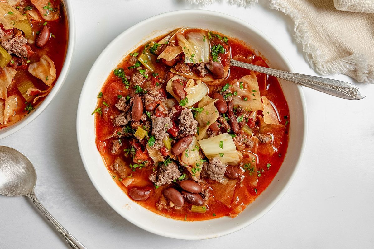 Taste Recipes Beef Cabbage Soup in white ceramic bowls with silver spoons and a linen napkin on a marble surface.