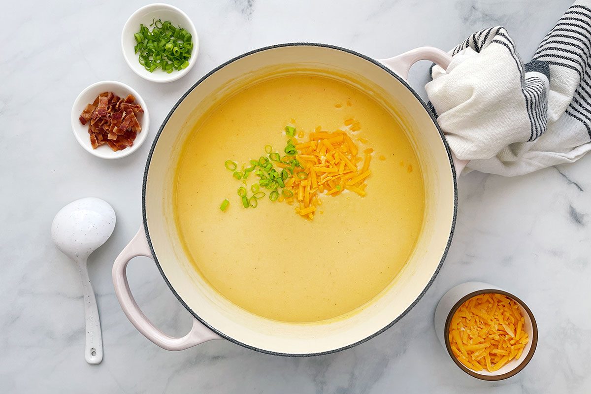 Overhead shot of Taste of Home's Beer Cheese Soup in a Dutch oven on a white marble background with bacon and green onions on the side