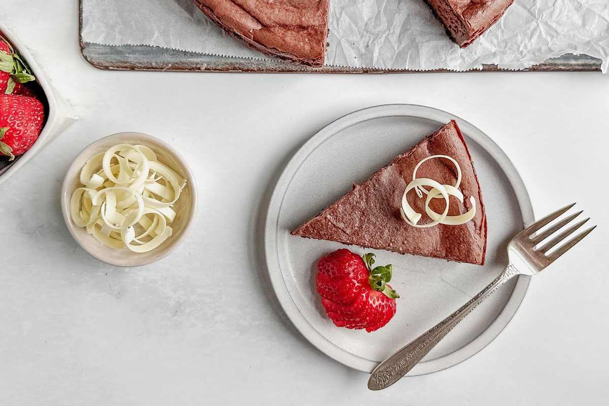 Taste Recipes chocolate cheesecake on a white plate with strawberries and white chocolate curls with a linen napkin on a marble surface