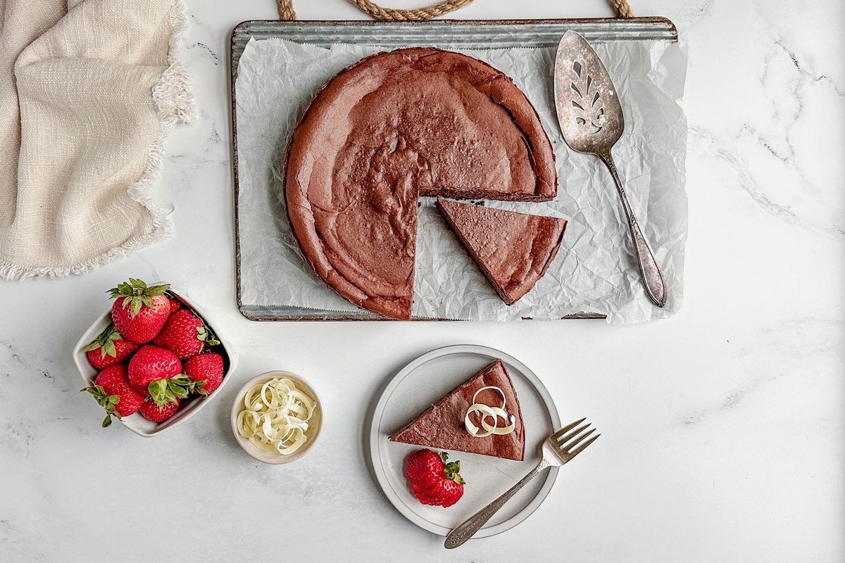 Taste Recipes chocolate cheesecake on a white plate with strawberries and white chocolate curls with a linen napkin on a marble surface.