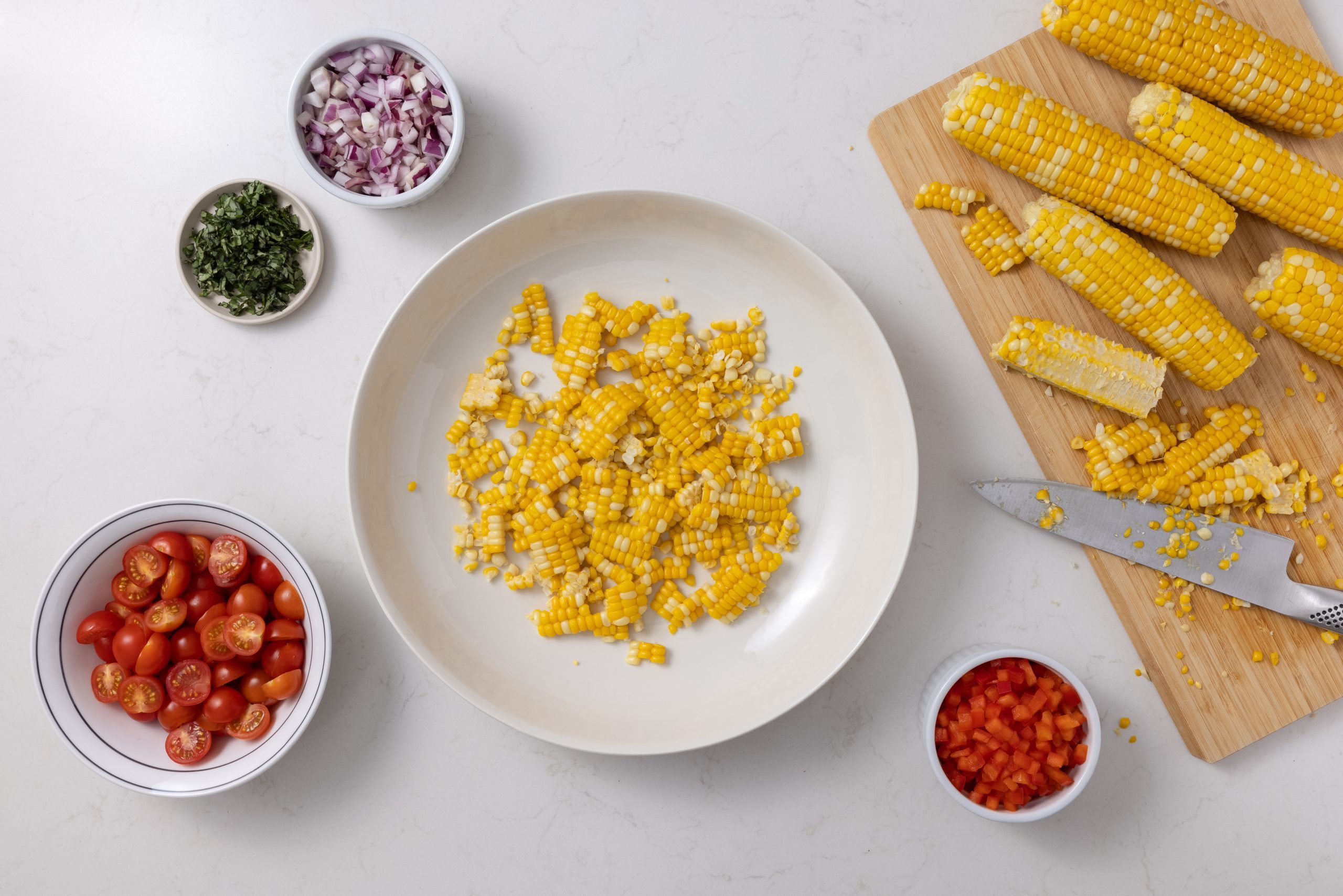 Corn being cut from the cob and mixed with other salad ingredients in large bowl.