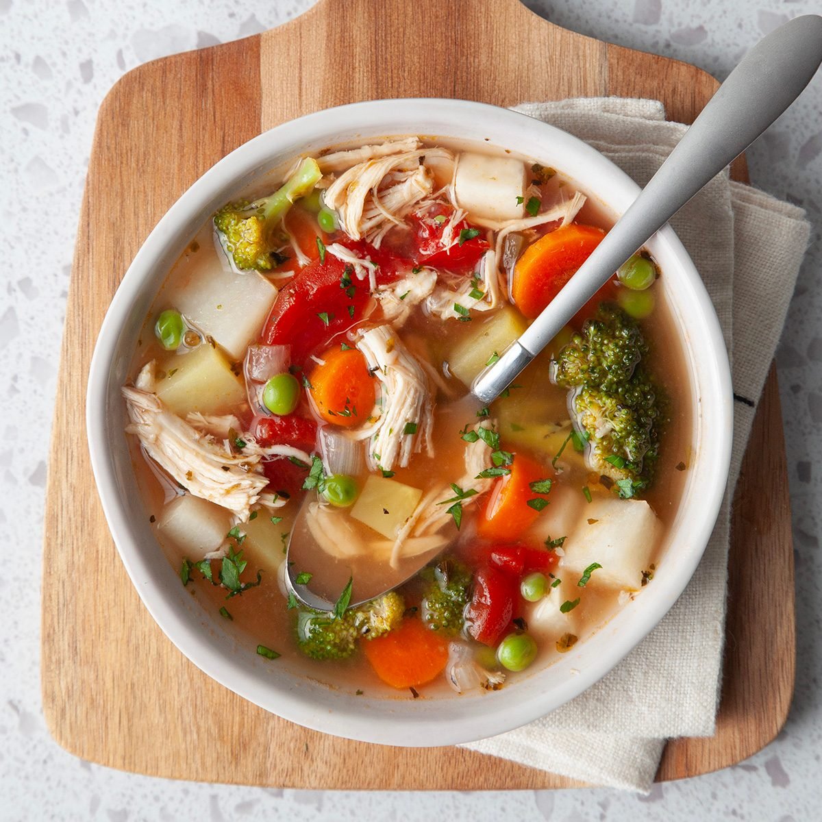 Single serving of Taste of Home Chicken Vegetable Soup in a white bowl on a wooden cutting board and a beige linen with a spoon