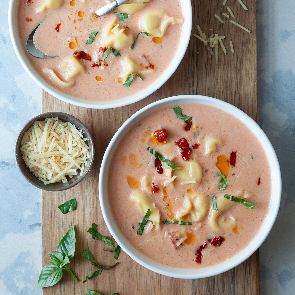 Two single servings of Taste of Home Tomato Tortellini Soup on a wooden cutting board, extra basil and parmesan on board