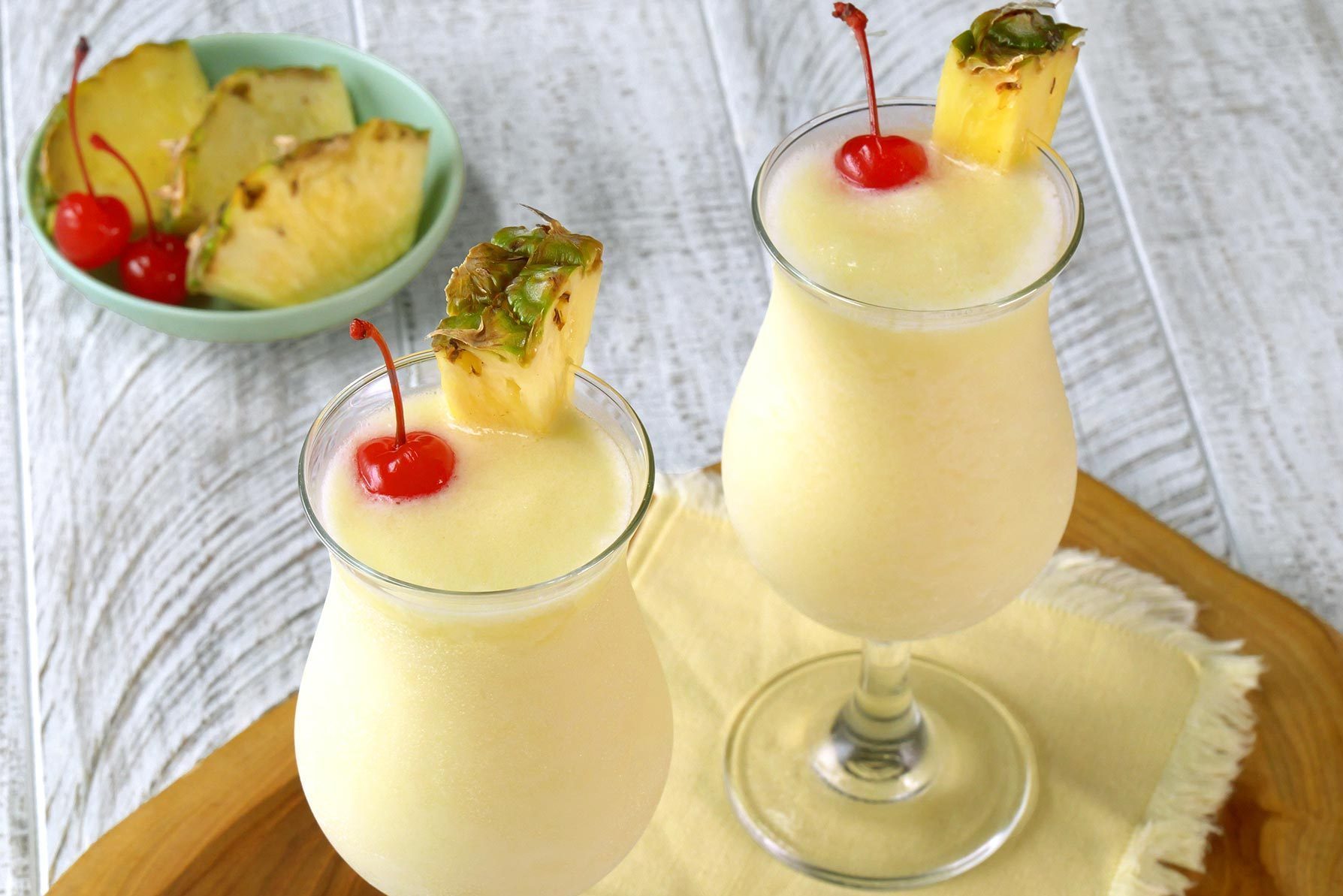 Two pina colada cocktails served in hurricane glasses, each garnished with a pineapple wedge and a maraschino cherry. 
