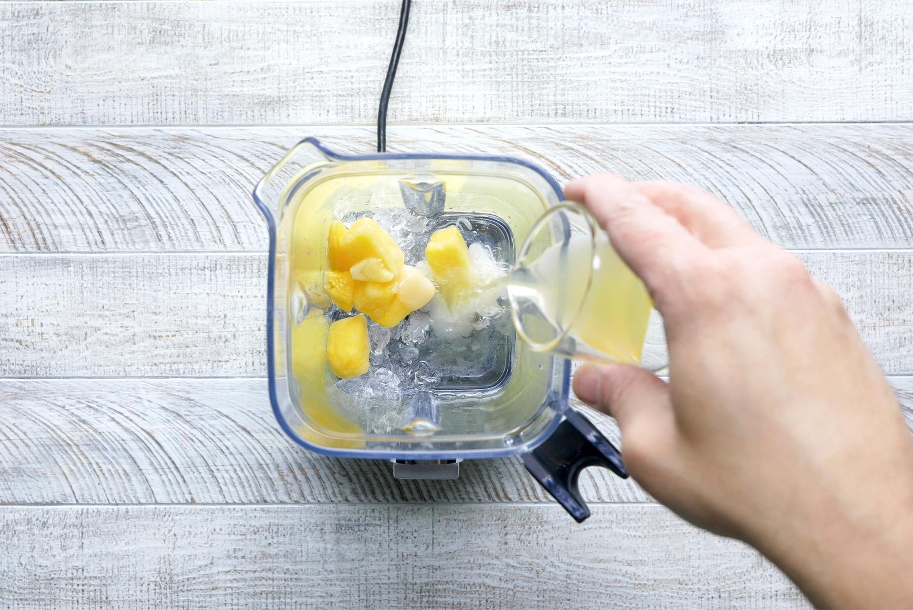 A hand pours a small glass of liquid into a blender containing ice cubes and chunks of mango, all set on a white wooden surface.