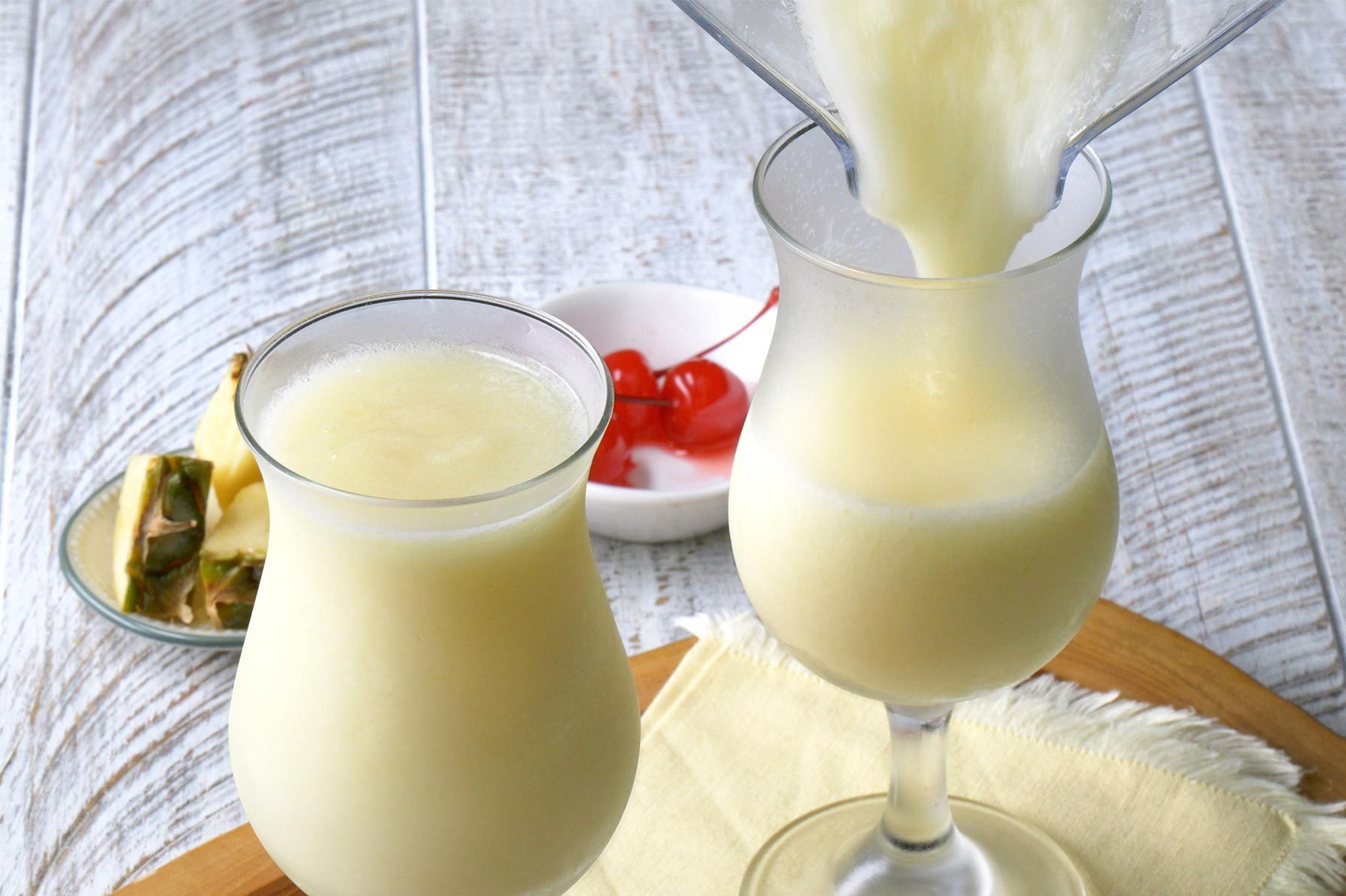 Two glasses of creamy piña colada are shown, one being poured from a blender. 