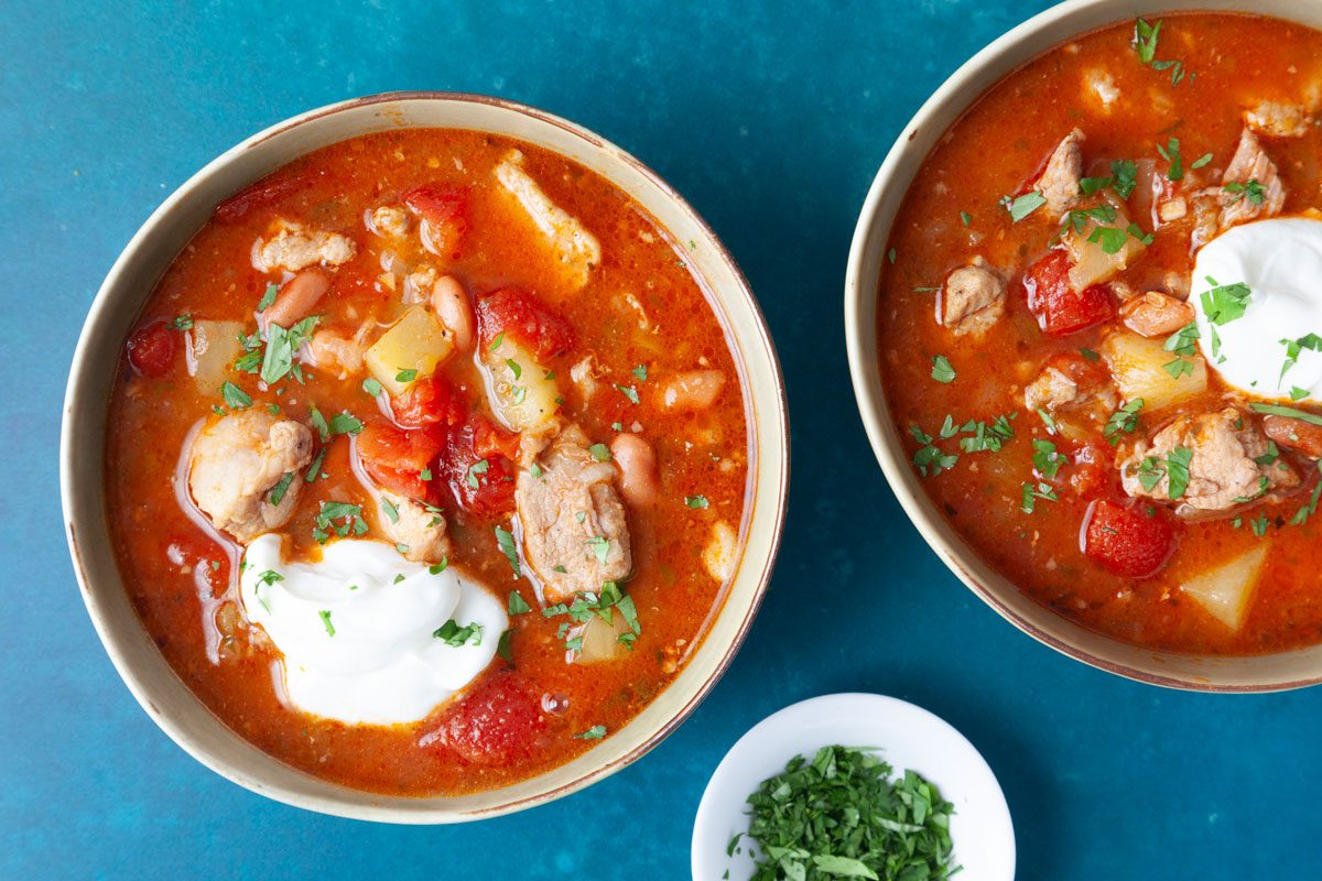 Two single servings of Taste of Home Green Chile Pork Stew in light green bowls on dark turquoise surface, garnished with sour cream and cilantro