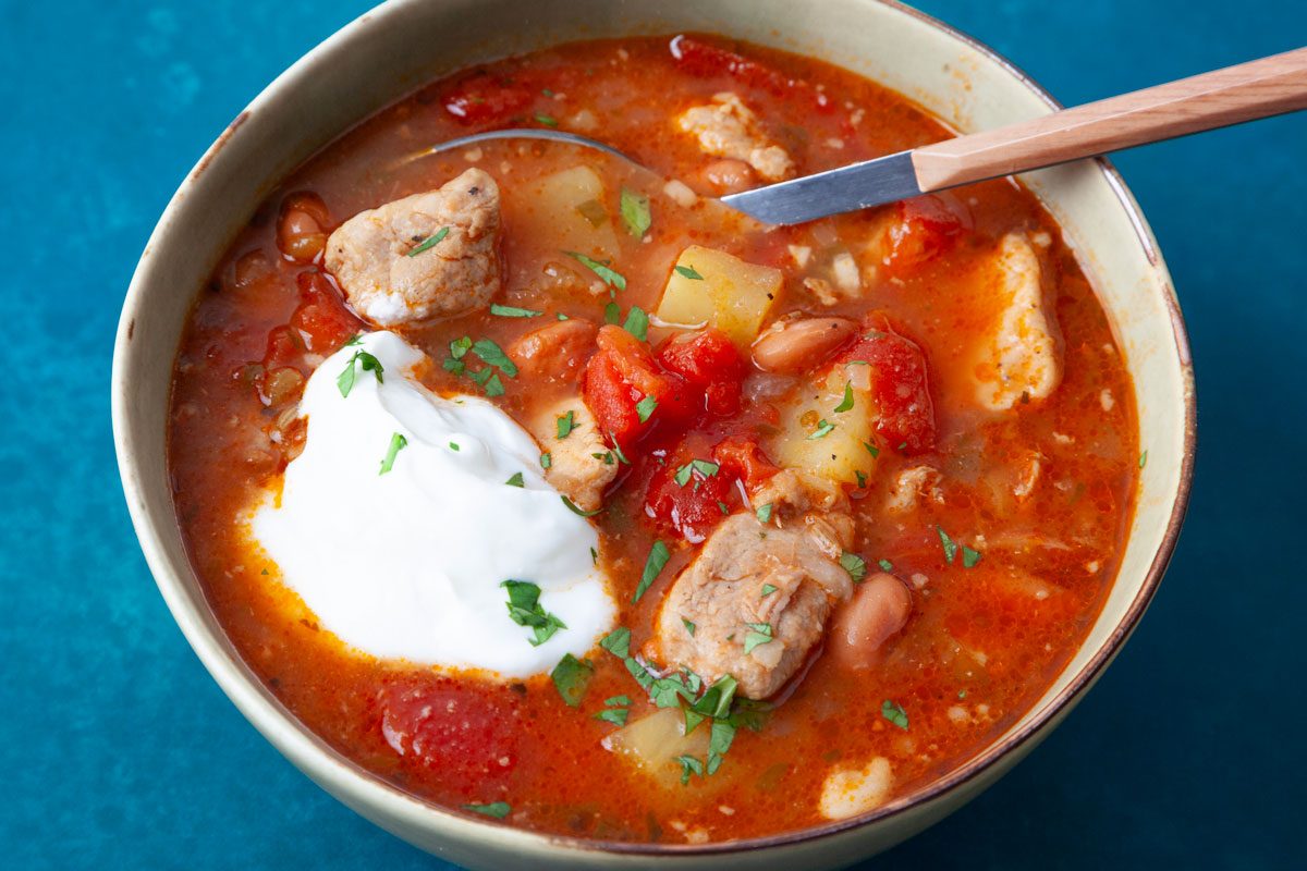 Single serving of Taste of Home Green Chile Pork Stew in light green bowl on dark turqoise surface, garnished with sour cream and cilantro