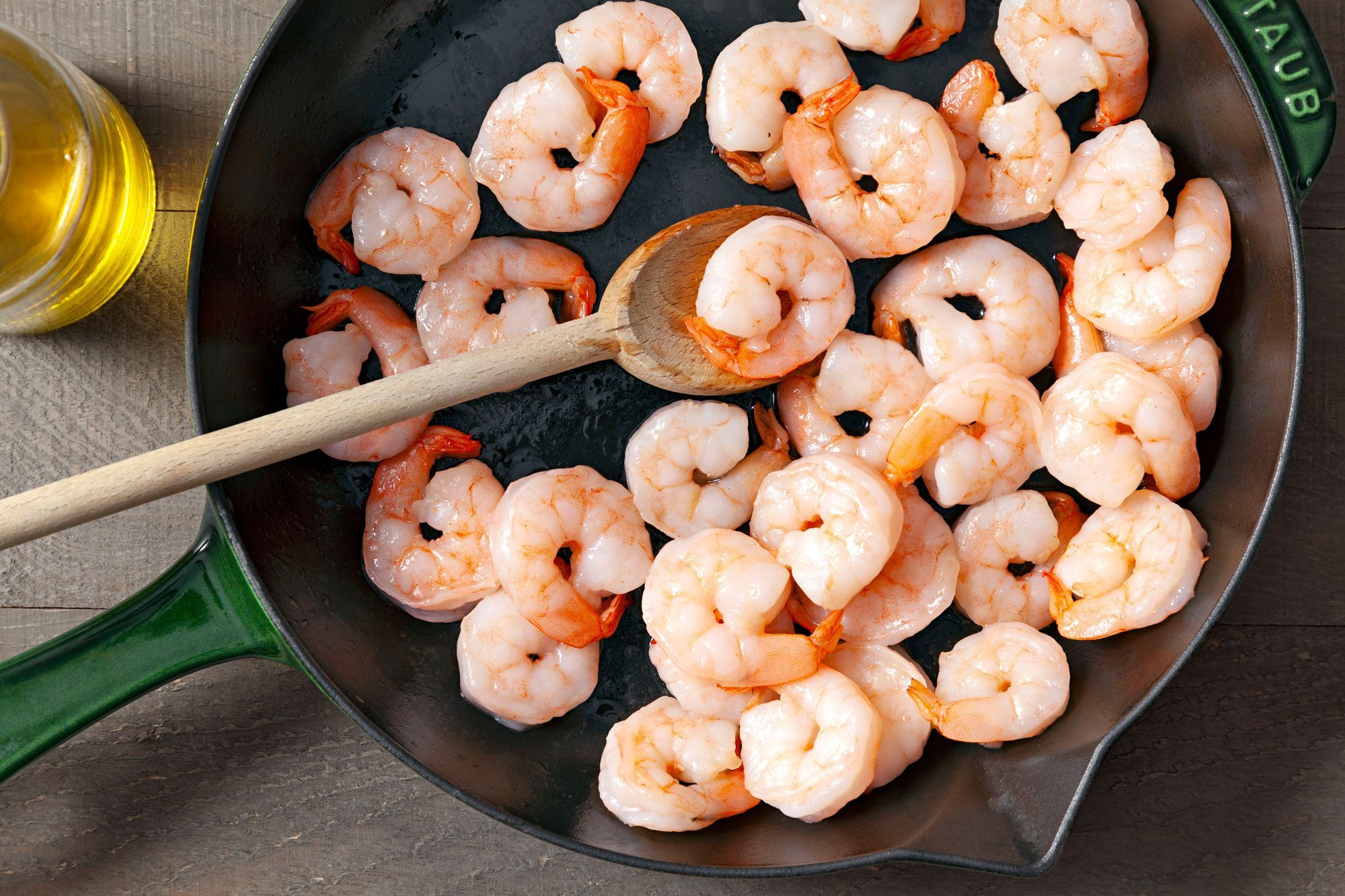 Overhead shot of a large cast-iron skillet; heat oil over medium-high heat; saute shrimp for 3 minutes; spoon; oil bottle; wooden background;