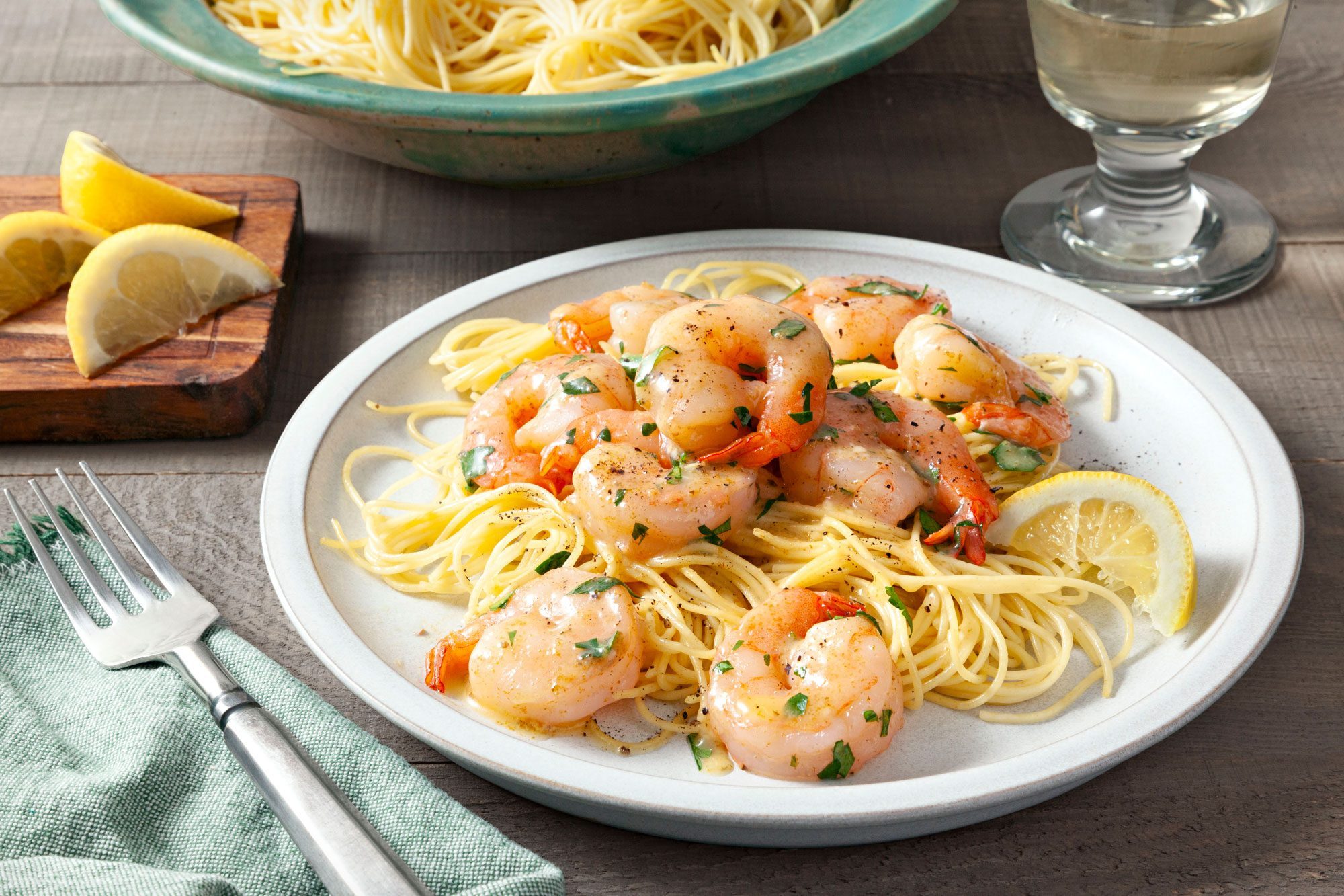 Table view shot of Garlic Lemon Shrimp; served in white plate with pasta; fork; napkin; lemon wedges; wood tray; glass of drink; wooden background;