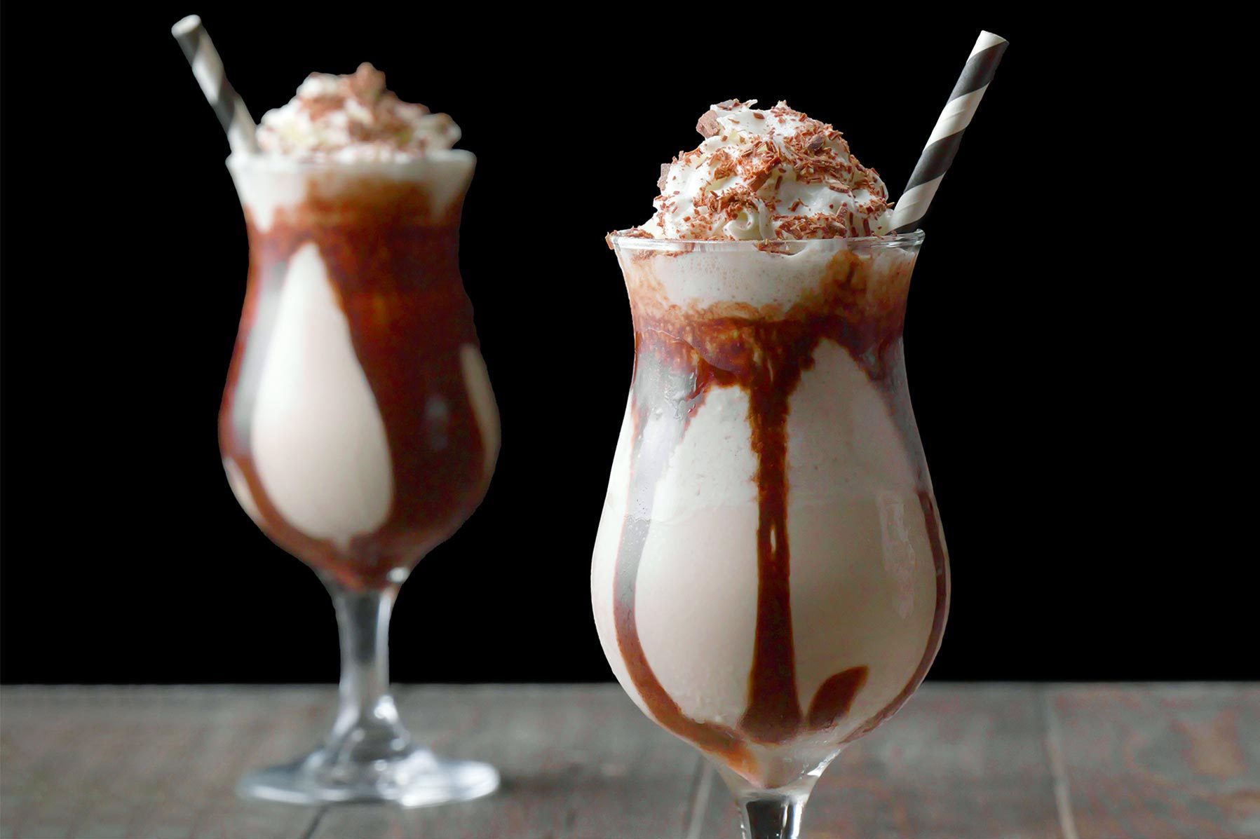 Two tall glasses of creamy chocolate milkshakes topped with whipped cream, shaved chocolate, and striped straws. 