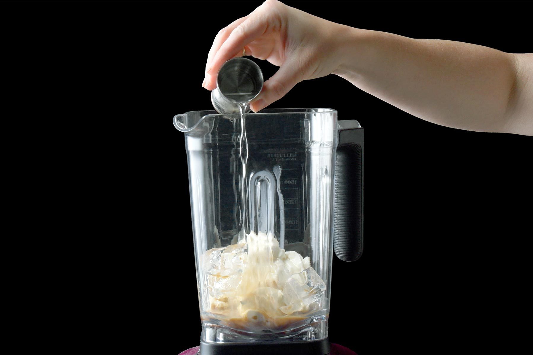 A hand is pouring a liquid from a small container into a blender jar filled with ice. The blender is clear and placed against a black background.