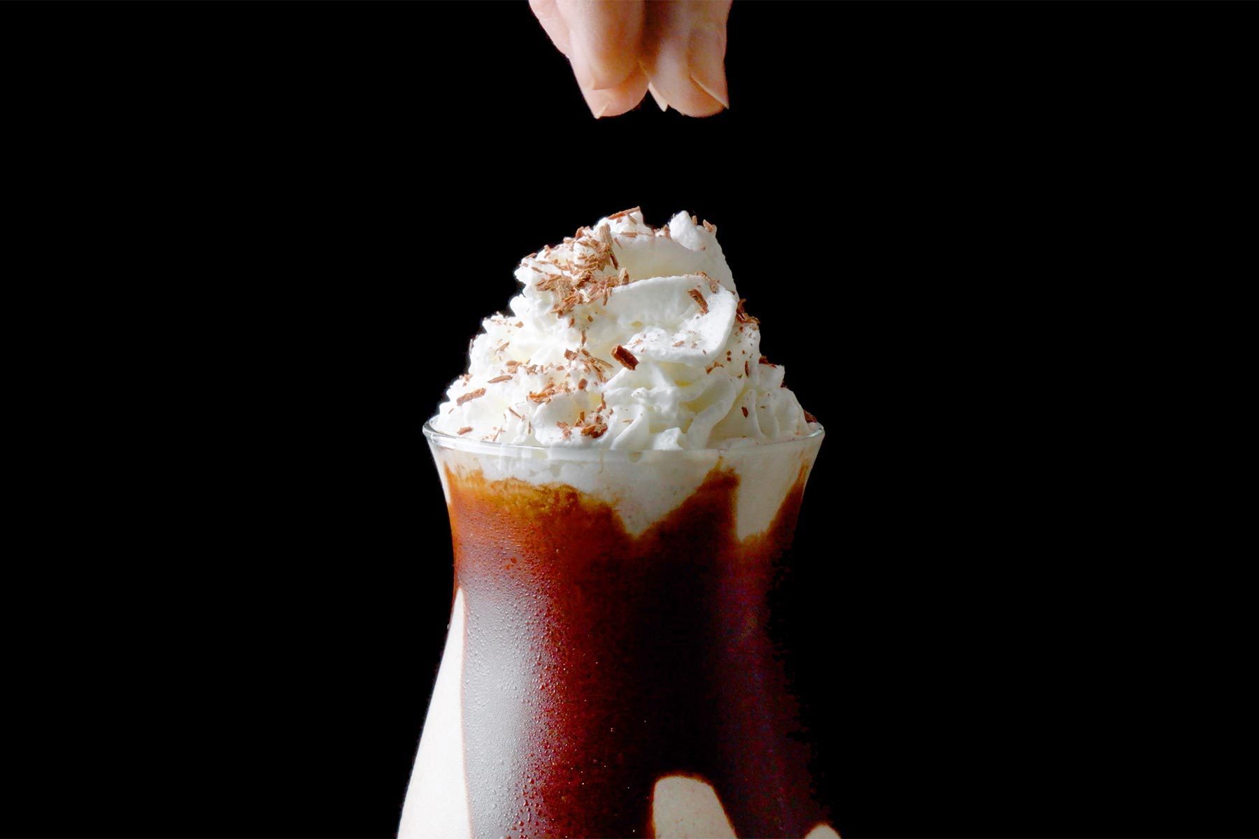 A hand is sprinkling chocolate shavings on top of a glass filled with a layered beverage.