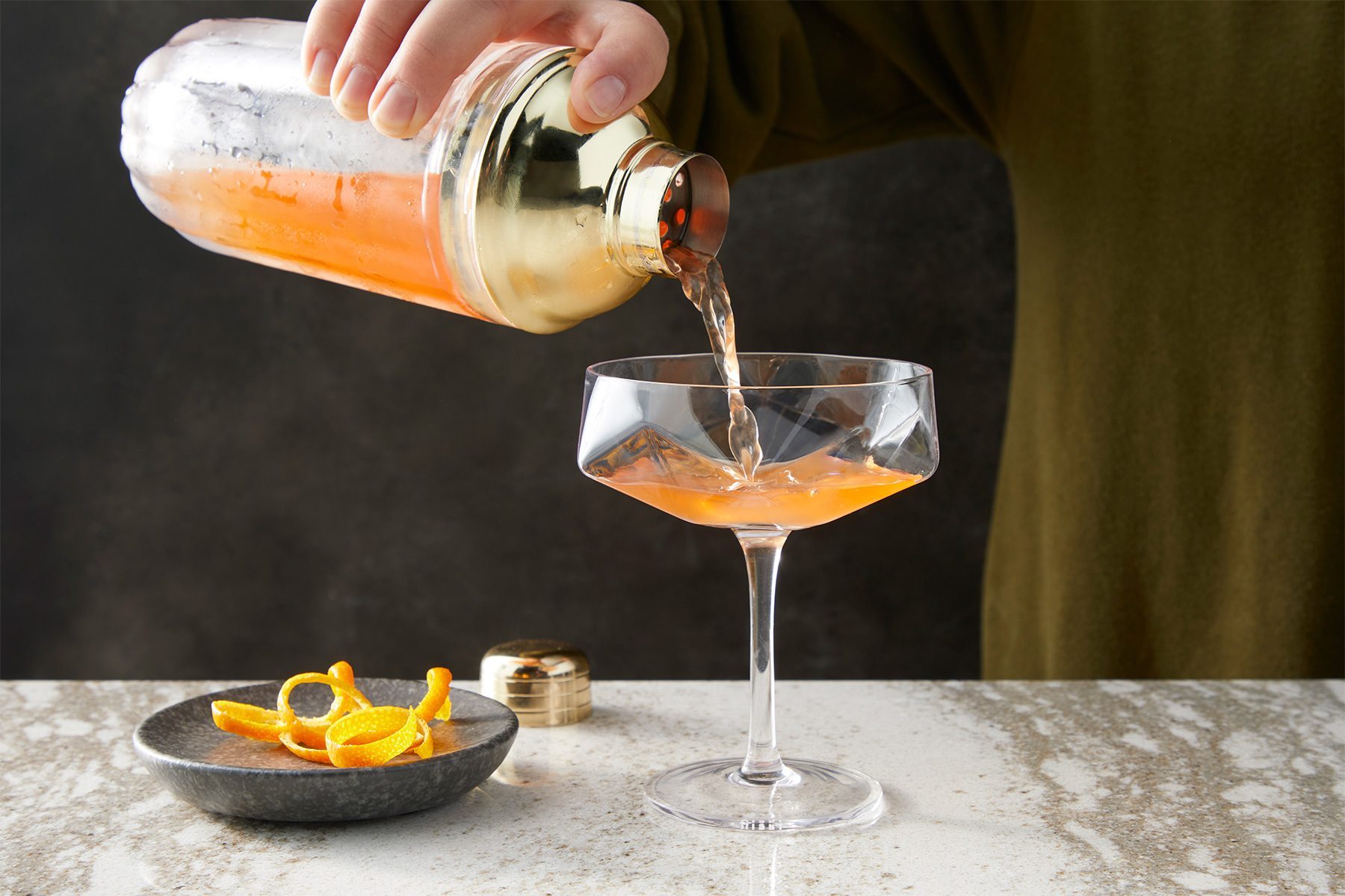 wide shot of paper plane drink strained into coupe glass; orange peel;