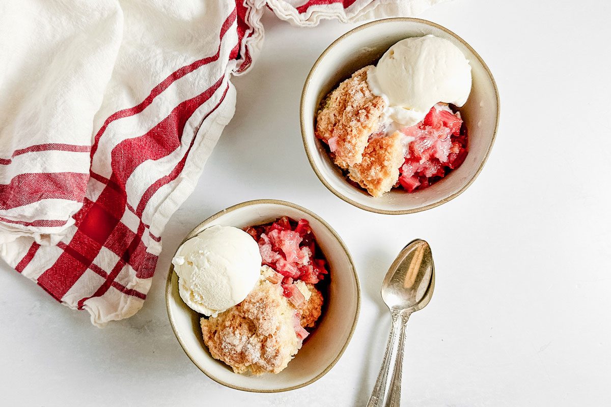 Taste of Home rhubarb cobbler and vanilla ice cream in bowls on a marble surface next to a red and white napkin.