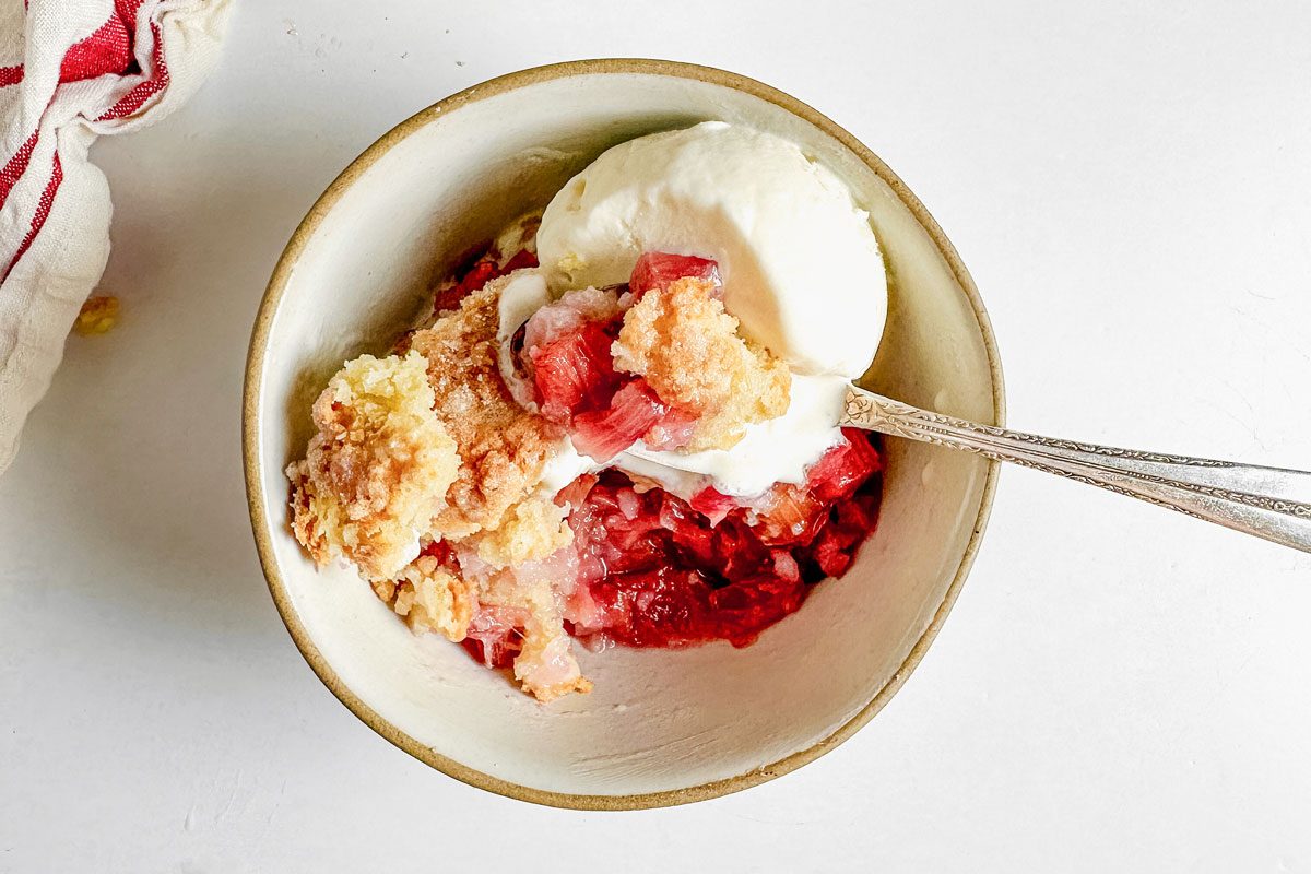 Taste of Home rhubarb cobbler and vanilla ice cream in a bowl on a marble surface next to a red and white napkin.