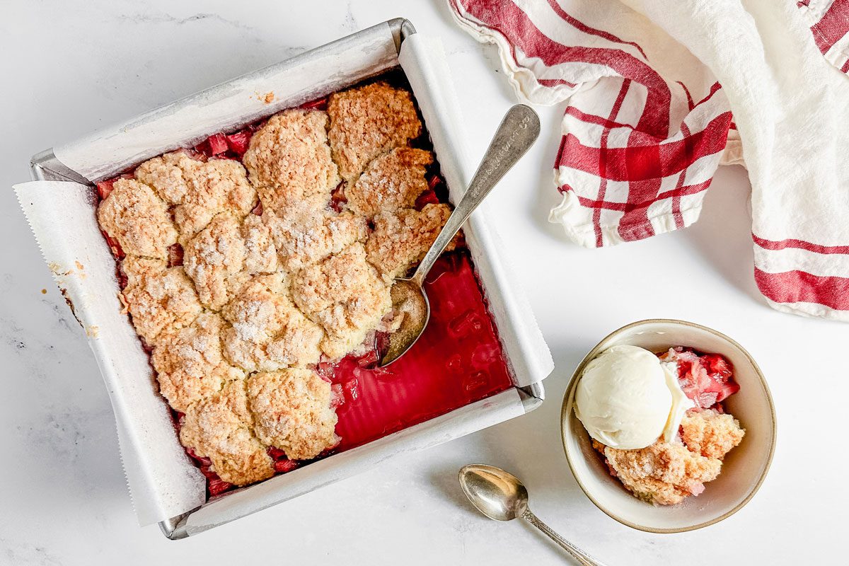 Taste of Home rhubarb cobbler baked in a metal pan on a marble surface next to a red and white napkin and a bowl of rhubarb cobbler with vanilla ice cream.