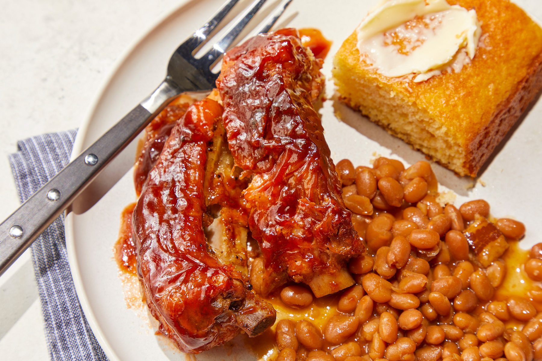 Close view shot of Slow-Cooker Baby Back Ribs; served on plate; with beans; cornbread and muffin; fork; napkin; marble background
