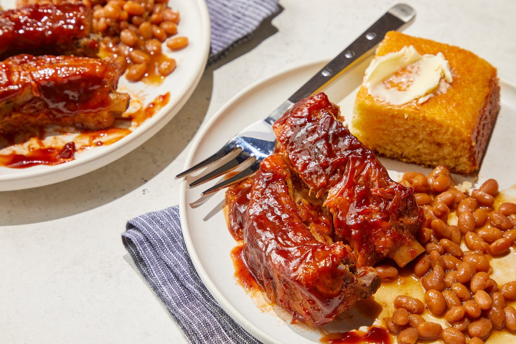 High angle view shot of Slow-Cooker Baby Back Ribs; served on plates; with beans; cornbread and muffin; fork; napkin; marble background