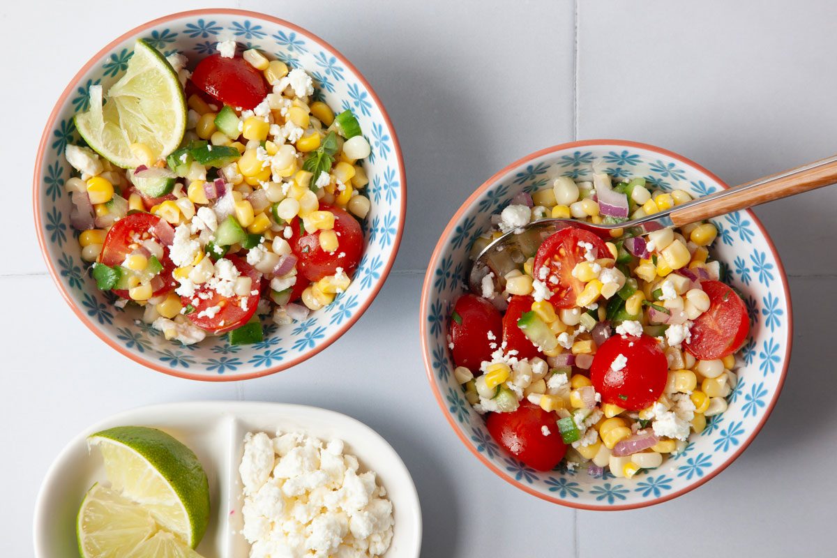 2 single servings of Taste of Home Summer Corn Salad in a blue and white bowls with feta and limes, 2 single servings in a blue and white bowls