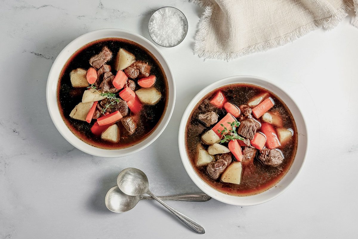 Taste of Home venison stew in white ceramic bowls on a marble surface next to a cloth napkin.