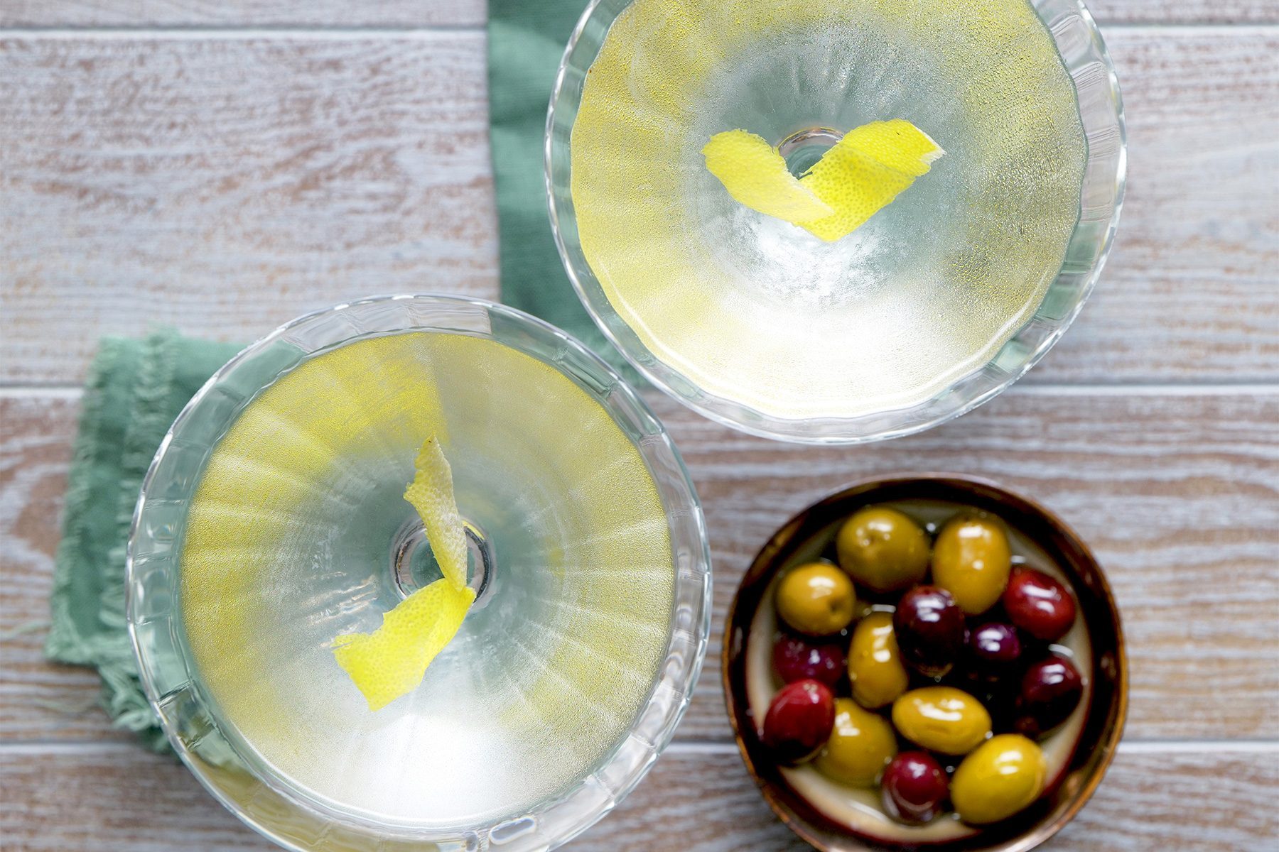 overhead shot; wooden background; Vesper Martini served in martini glasses with lemon peel inside it, over kitchen towel; served with olives;