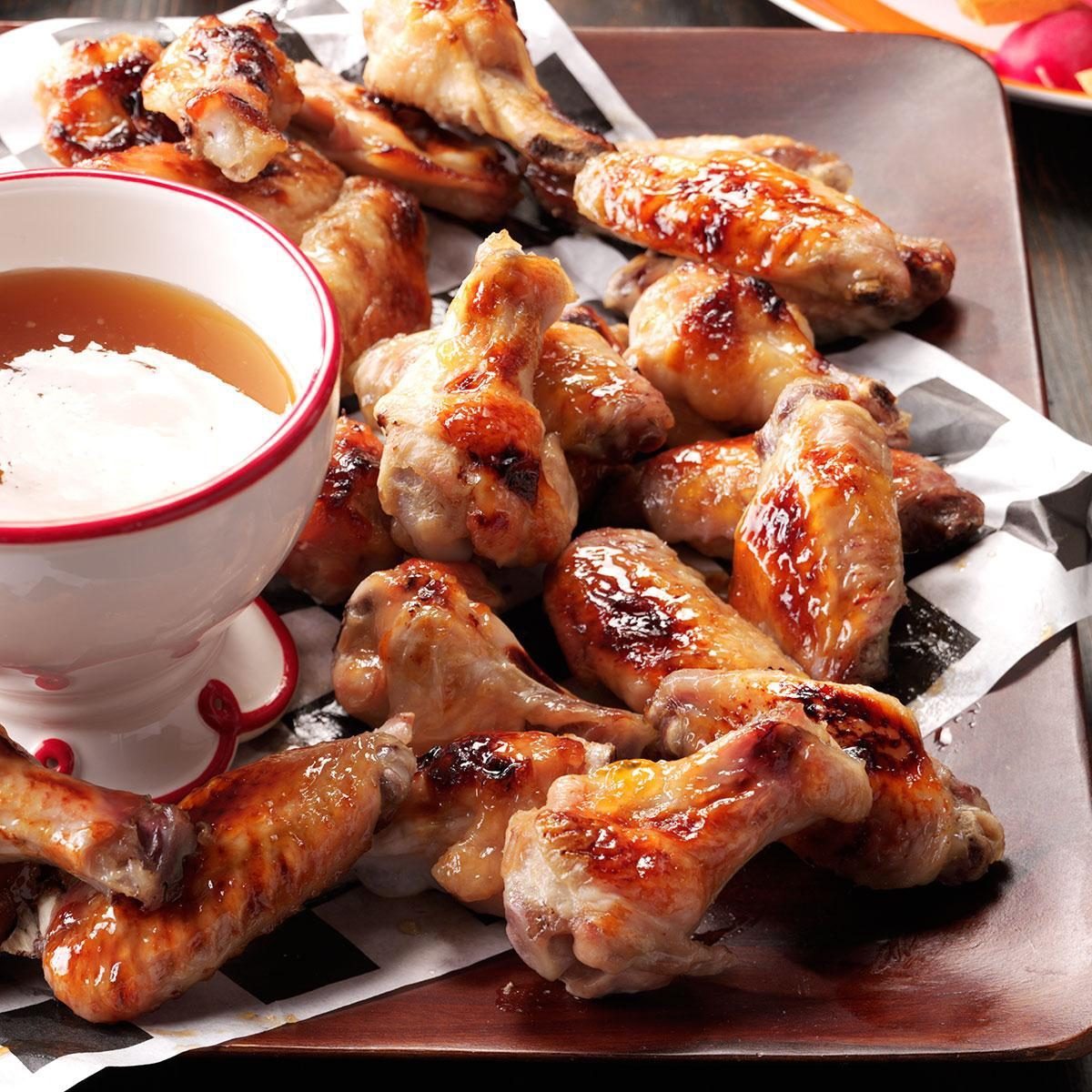 A platter of glazed chicken wings is served on a rectangular tray lined with paper. A small white bowl filled with dipping sauce sits on the tray, adding an appetizing touch to the dish.