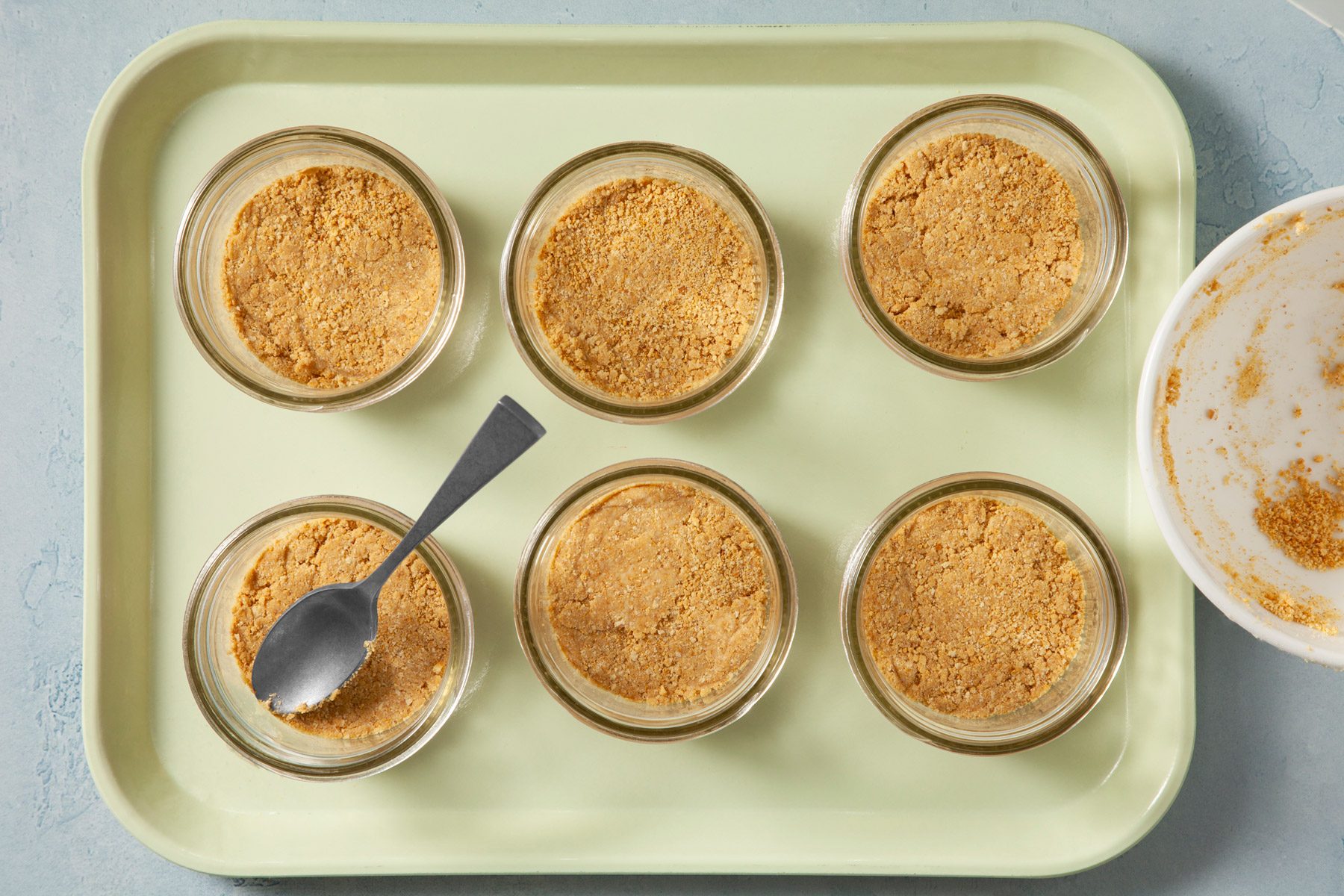 Graham cracker crumbs mixture divided and layered at the bottom of 6 cups. One cup has a spoon in it.