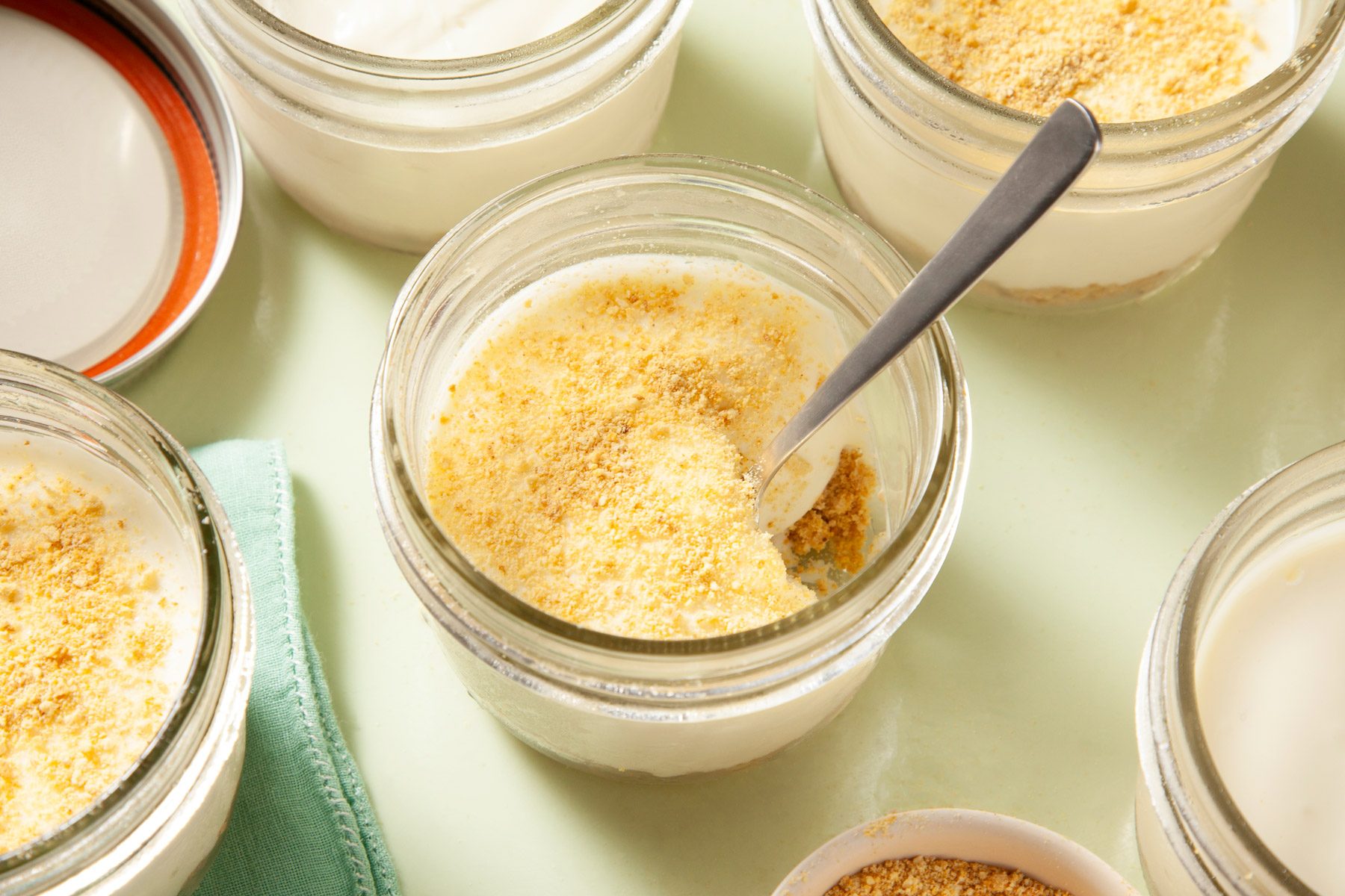 Glass jar containing cheesecake with a spoon in it. It is surrounded by other partially visible glass jars.