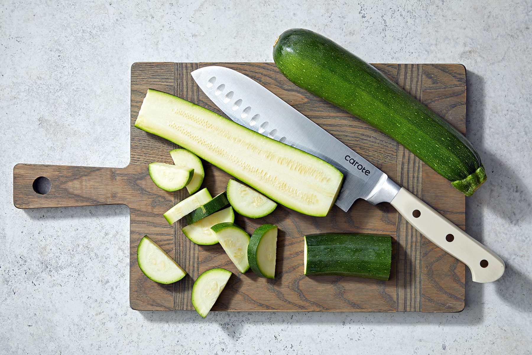 A wooden cutting board holds a partially chopped zucchini. There is a knife with a white handle and a whole zucchini placed next to it. Some zucchini pieces are sliced and arranged on the board. The background surface is light-colored.