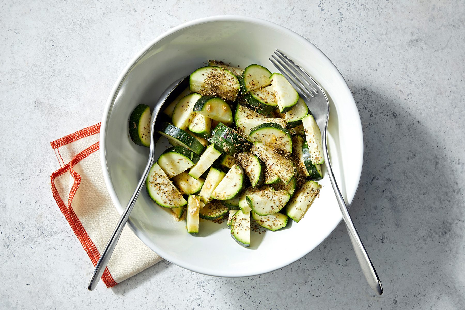 A bowl of sliced zucchini seasoned with spices, accompanied by forks. The bowl is placed on a lightly textured surface with a red-edged white napkin beside it.