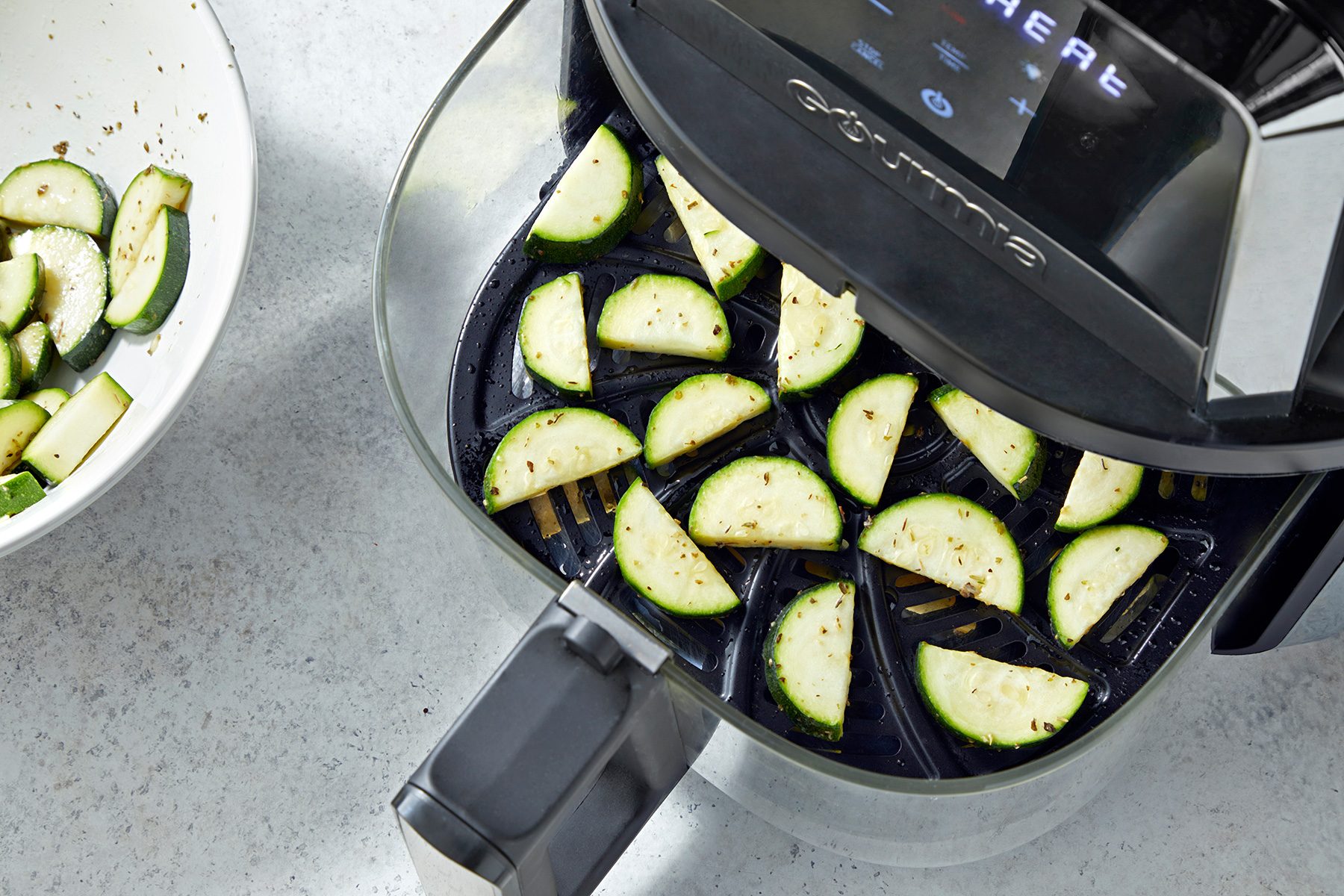 An air fryer is filled with zucchini slices seasoned with herbs and spices. A bowl on the side also contains seasoned zucchini pieces, ready to be air-fried. The appliances and food are placed on a light-colored kitchen countertop.