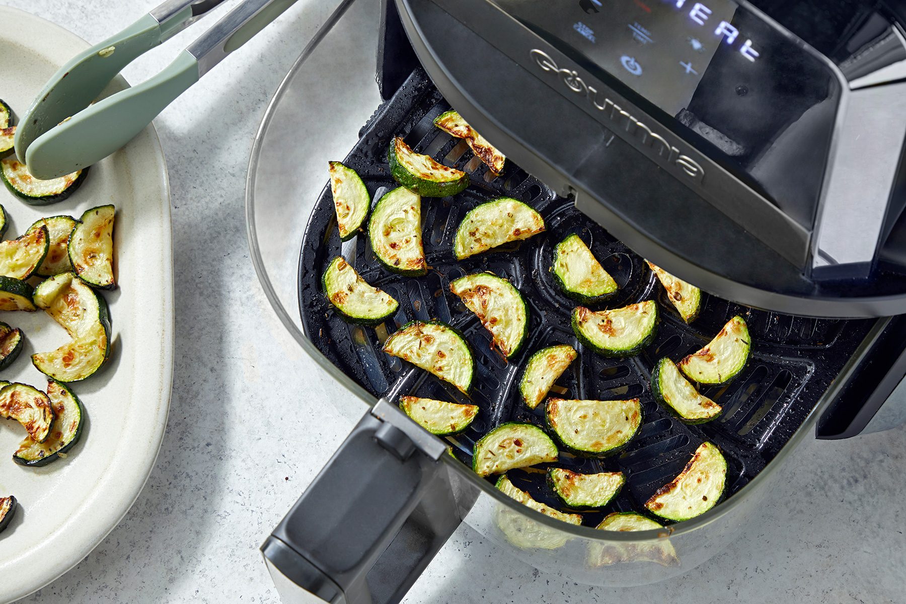 A top view of an air fryer with slices of zucchini inside, some cooked and some raw. A pair of tongs is seen next to the air fryer. A white plate with more slices of zucchini is nearby on a light-colored kitchen countertop.