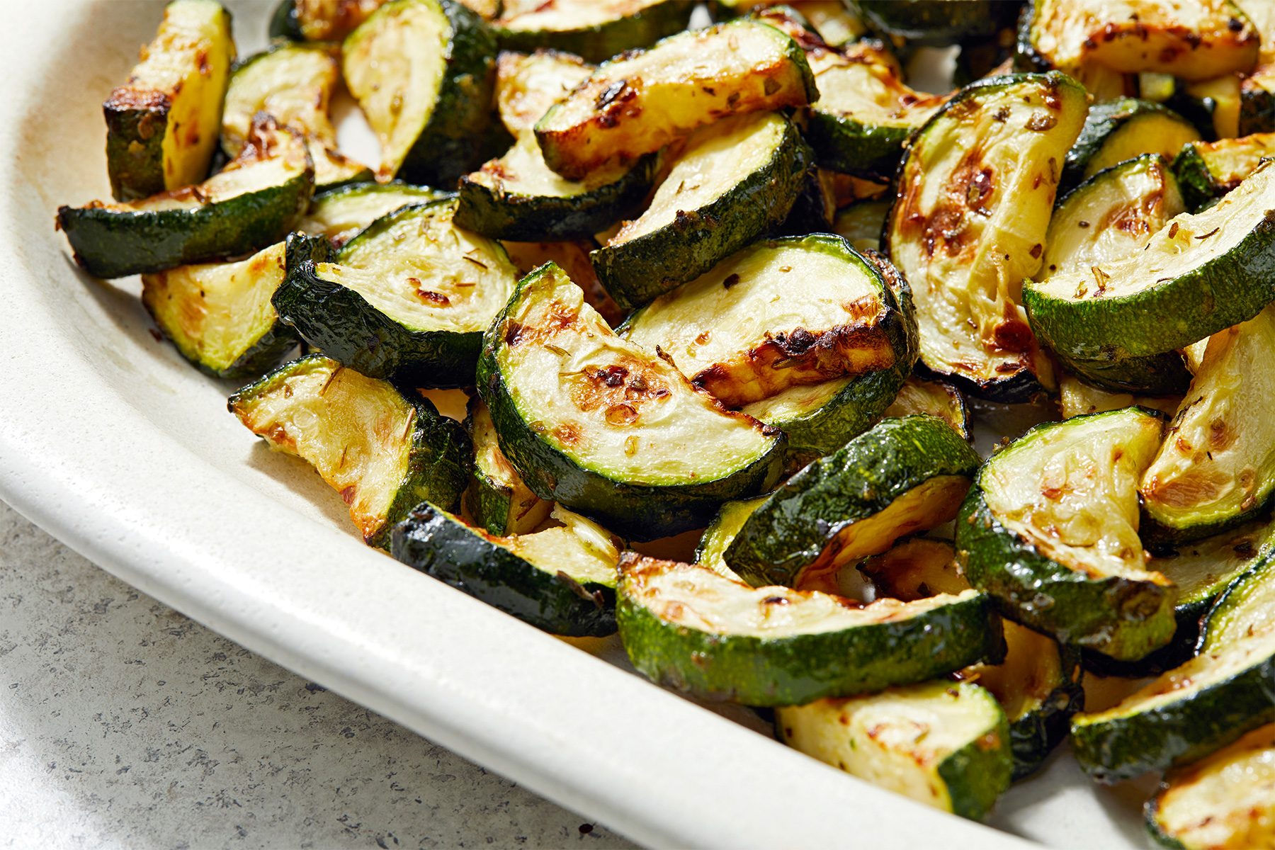 A close-up of a platter filled with roasted zucchini slices. The zucchini pieces are lightly browned and seasoned, giving them a flavorful appearance. The platter is set on a light-colored surface.