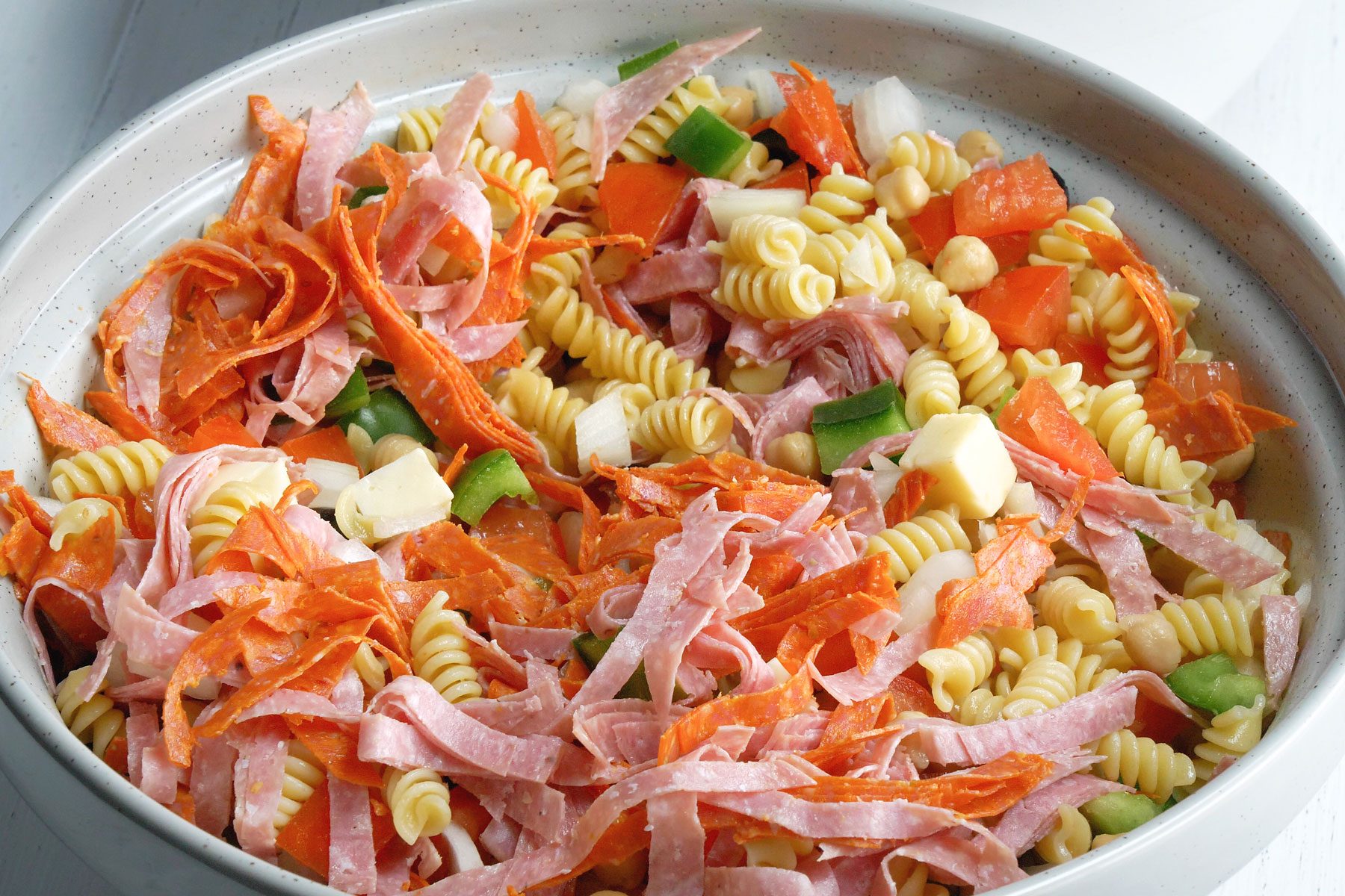 Large bowl containing pasta with the tomatoes, onions, green peppers, chickpeas, salami, pepperoni, provolone and olives.