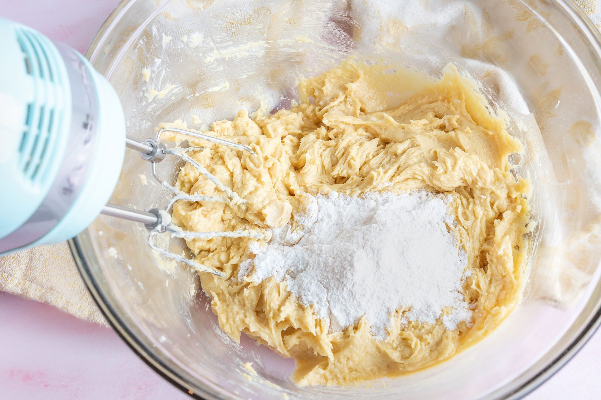 Overhead shot of a large bowl; cream butter and sugars until light and fluffy; beat pudding mix; eggs and vanilla; in another bowl, whisk flour; baking soda and salt; blender; kitchen towel; pink texture surface;