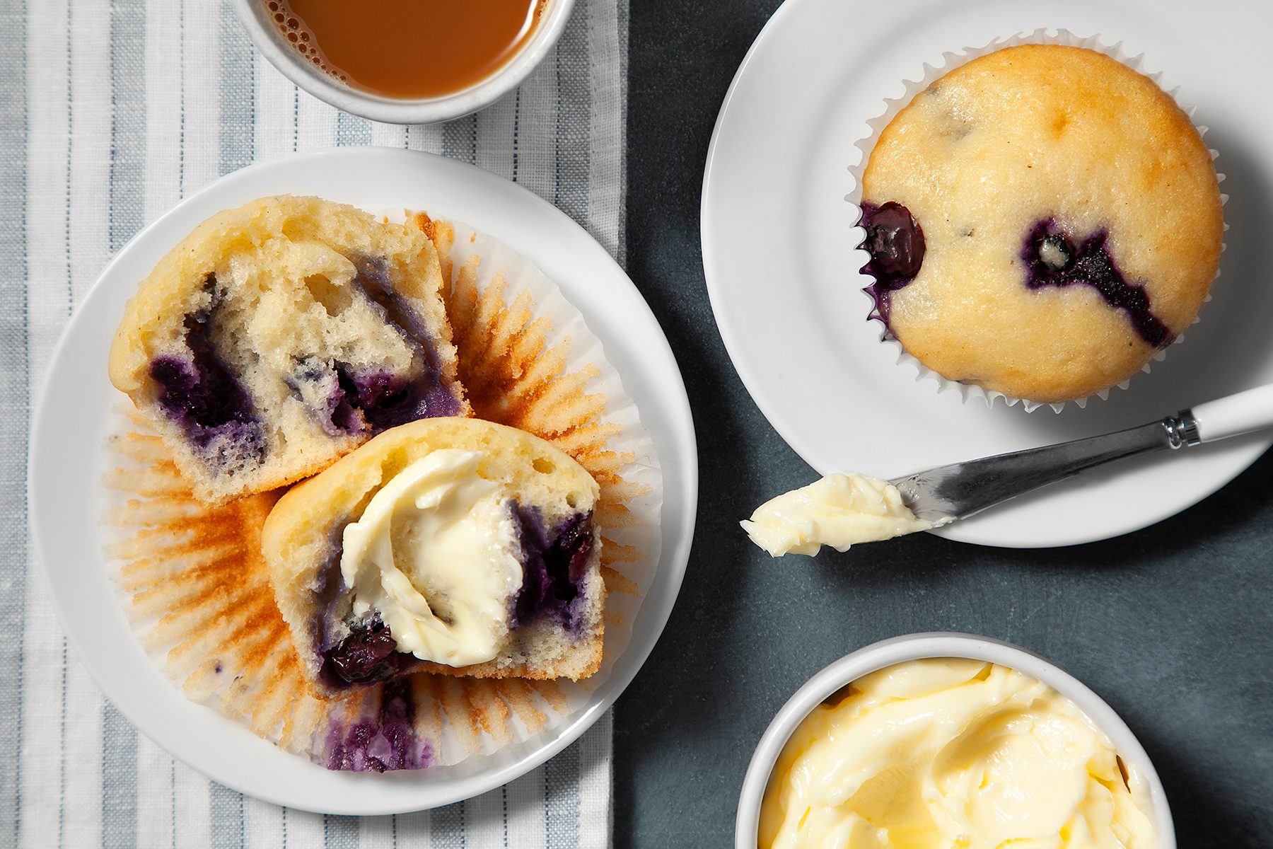 A blueberry muffin, cut in half and topped with butter, sits on a striped napkin with a cup of coffee or tea nearby. 