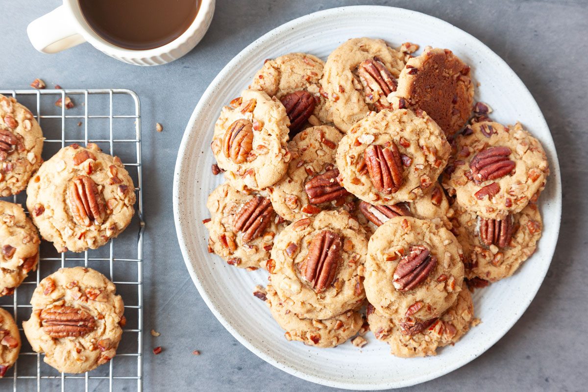 Full yield of Taste of Home Butter Pecan Cookies on a cooling rack and a white plate, cup of coffee peaking in