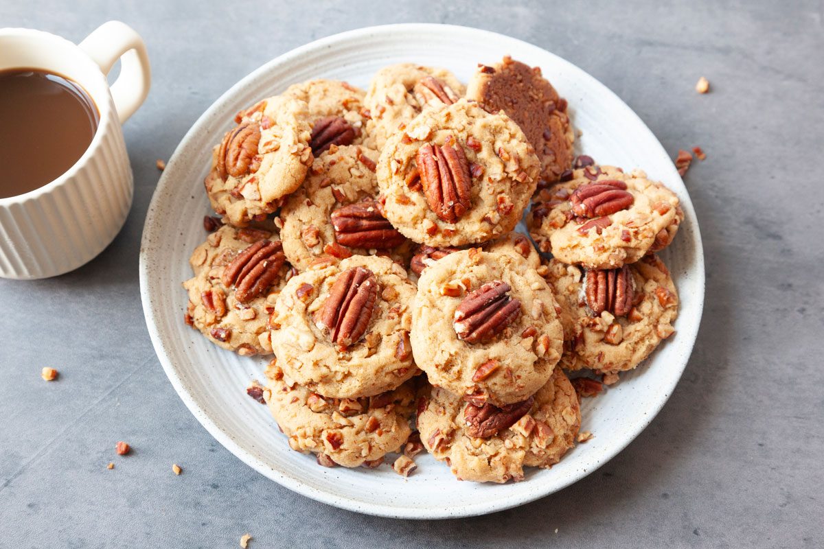 Full yield of Taste of Home Butter Pecan Cookies on a white plate, cup of coffee peaking in