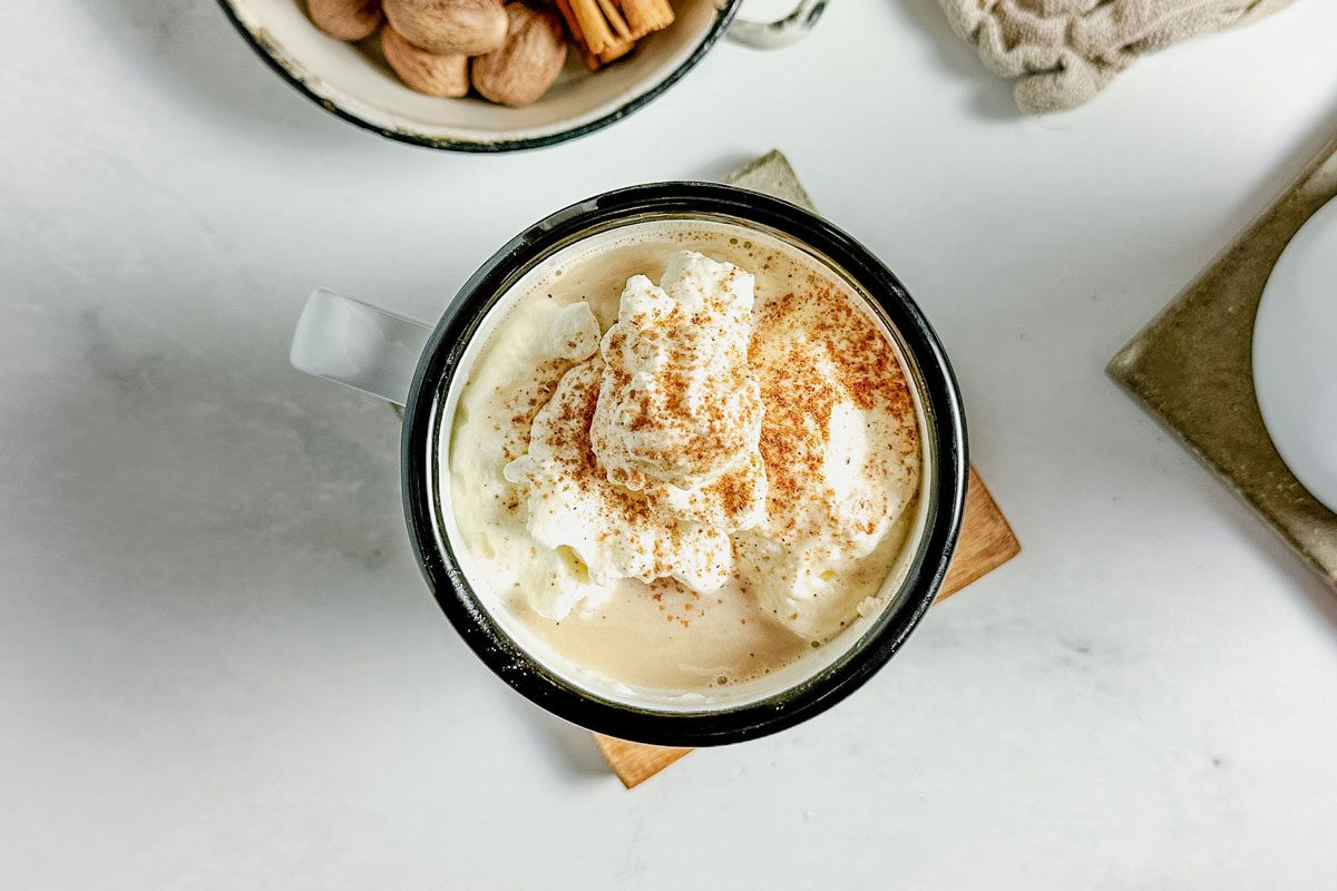 Taste Recipes Chai Tea Latte in enamel mugs on wooden coasters with an enamel dish filled with cinnamon sticks and whole nutmeg on a marble surface.