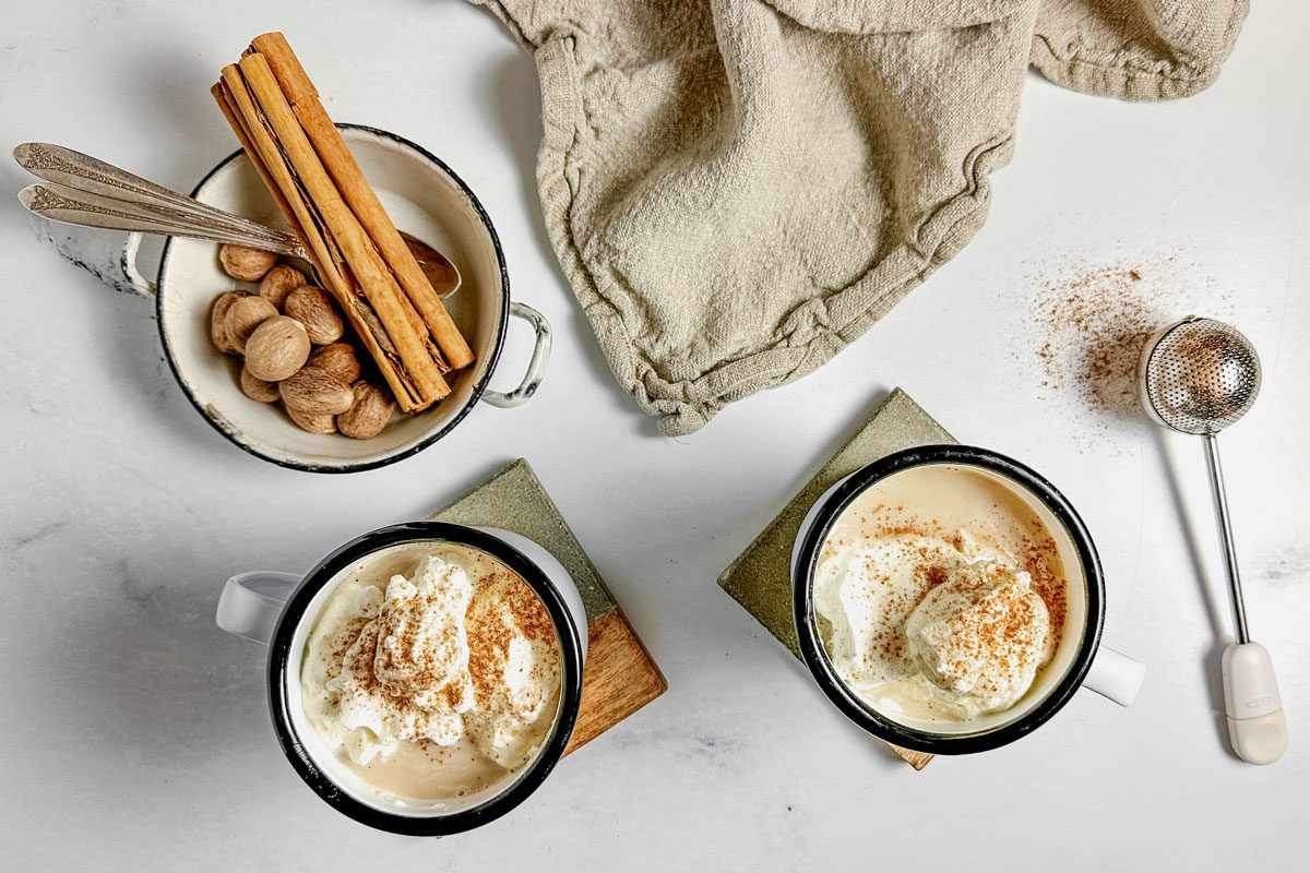 Taste Recipes Chai Tea Latte in enamel mugs on wooden coasters with an enamel dish filled with cinnamon sticks and whole nutmeg on a marble surface.