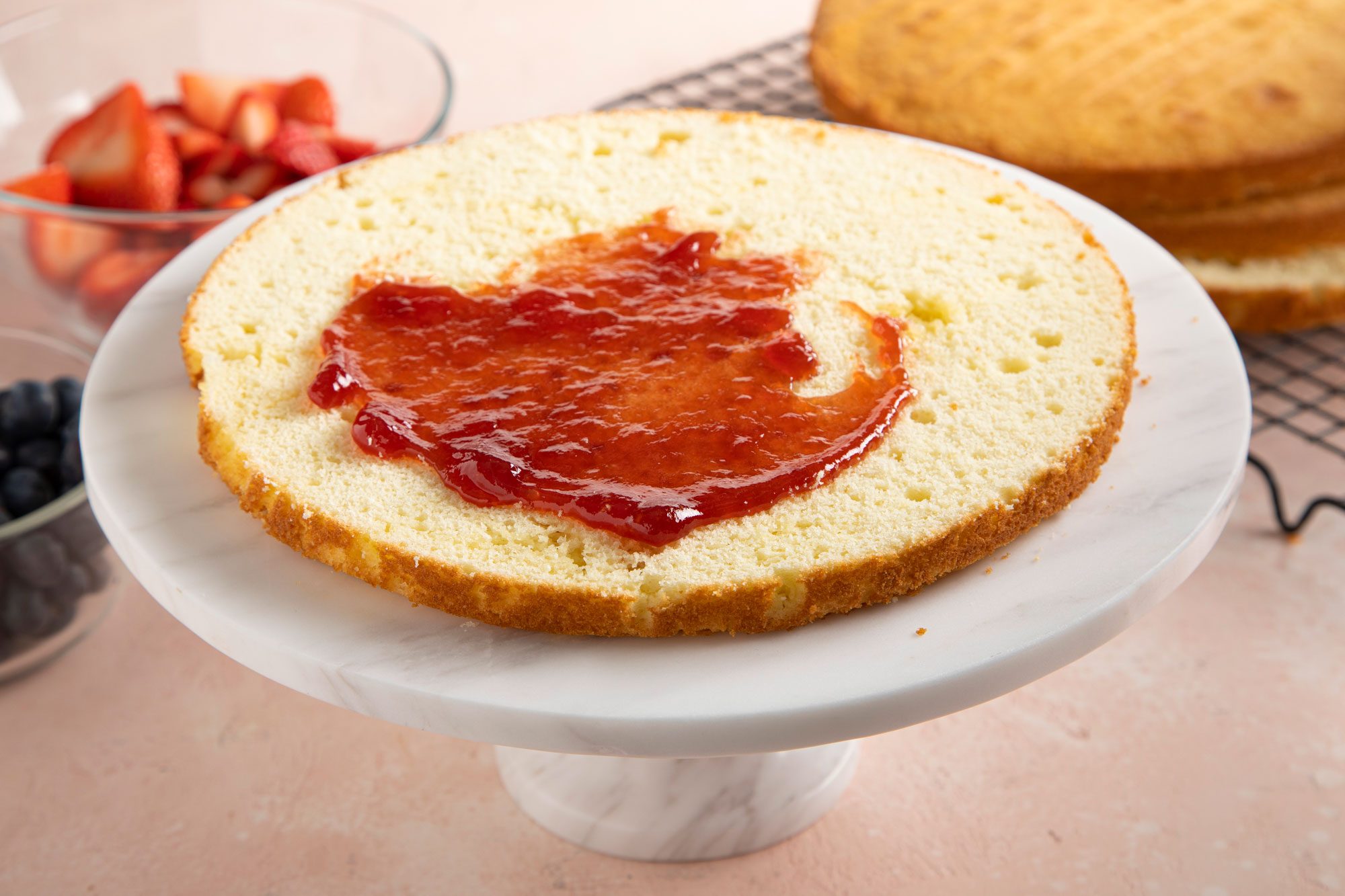 A round cake layer on a white marble cake stand, with a spread of strawberry jam on top.