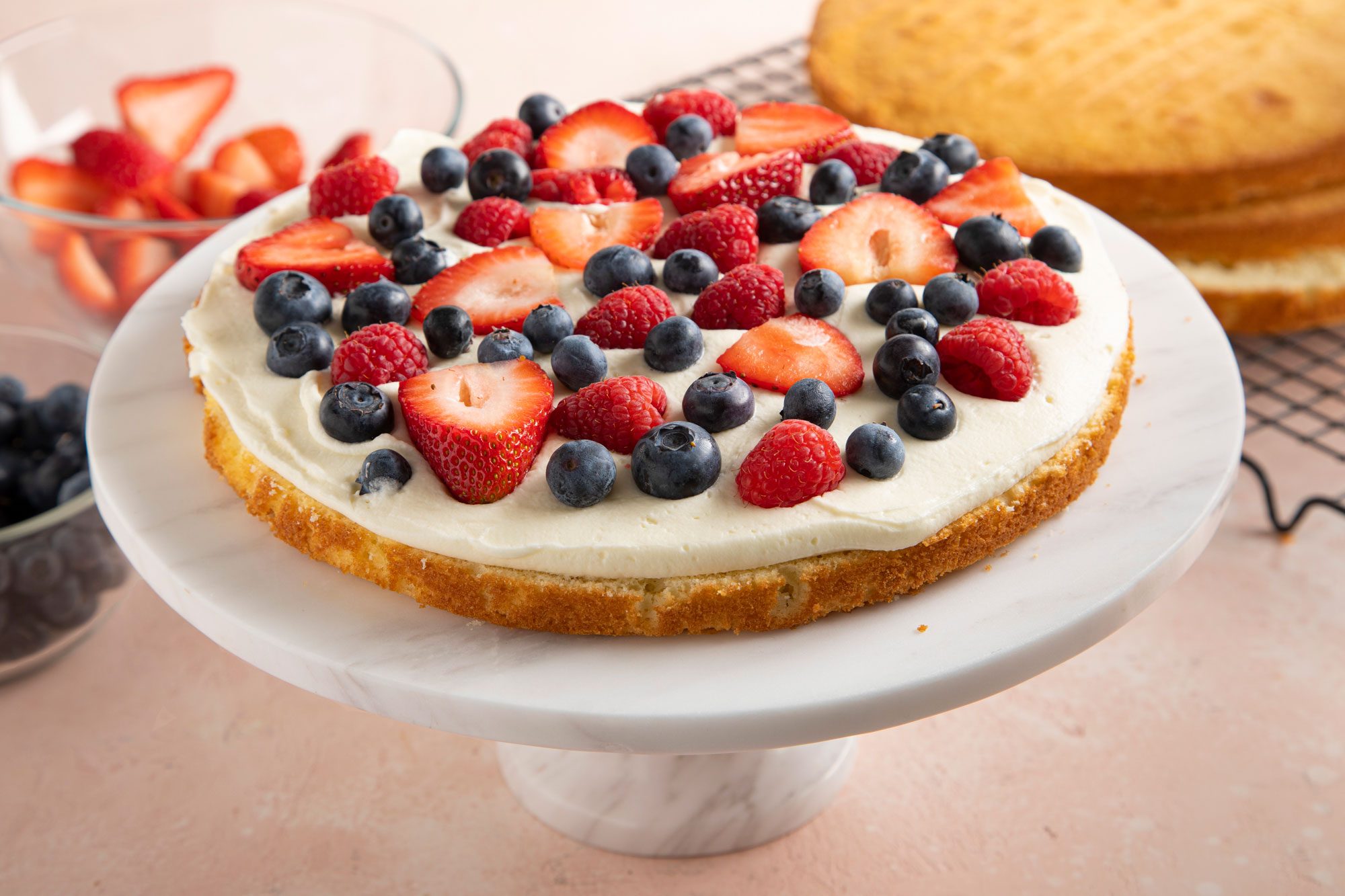 A round cake layer on a white marble cake stand, with a spread of frosting, strawberries and blueberries on top.