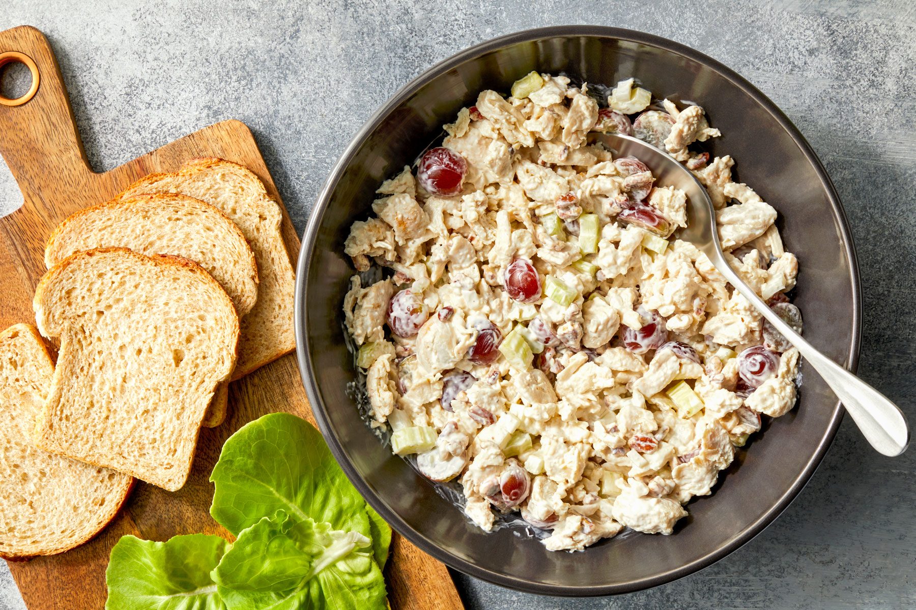 Chicken salad in a large bowl. Bread and lettuce are next to it on a wooden chopping board.