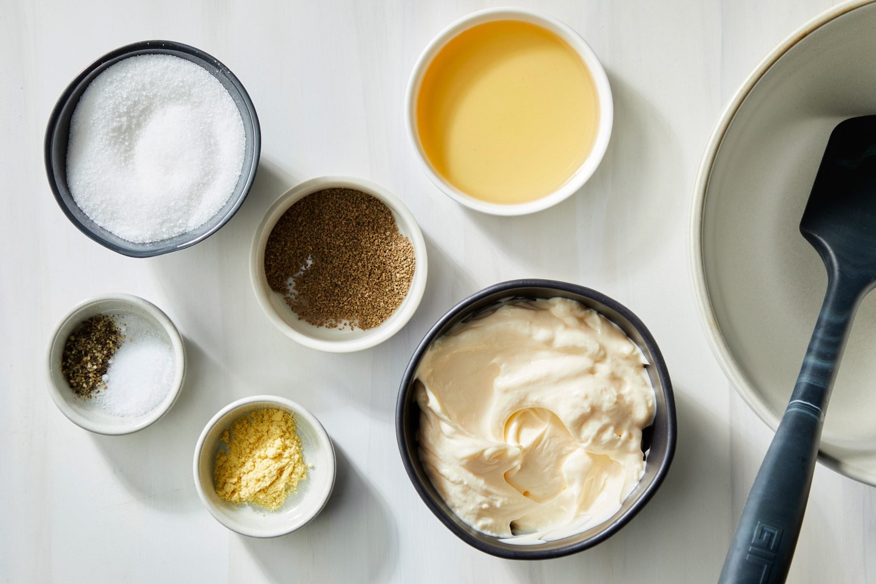 A collection of small bowls containing various ingredients, including mayonnaise, salt, mustard powder and spices arranged on a white surface. A spatula and a beige plate are also visible.